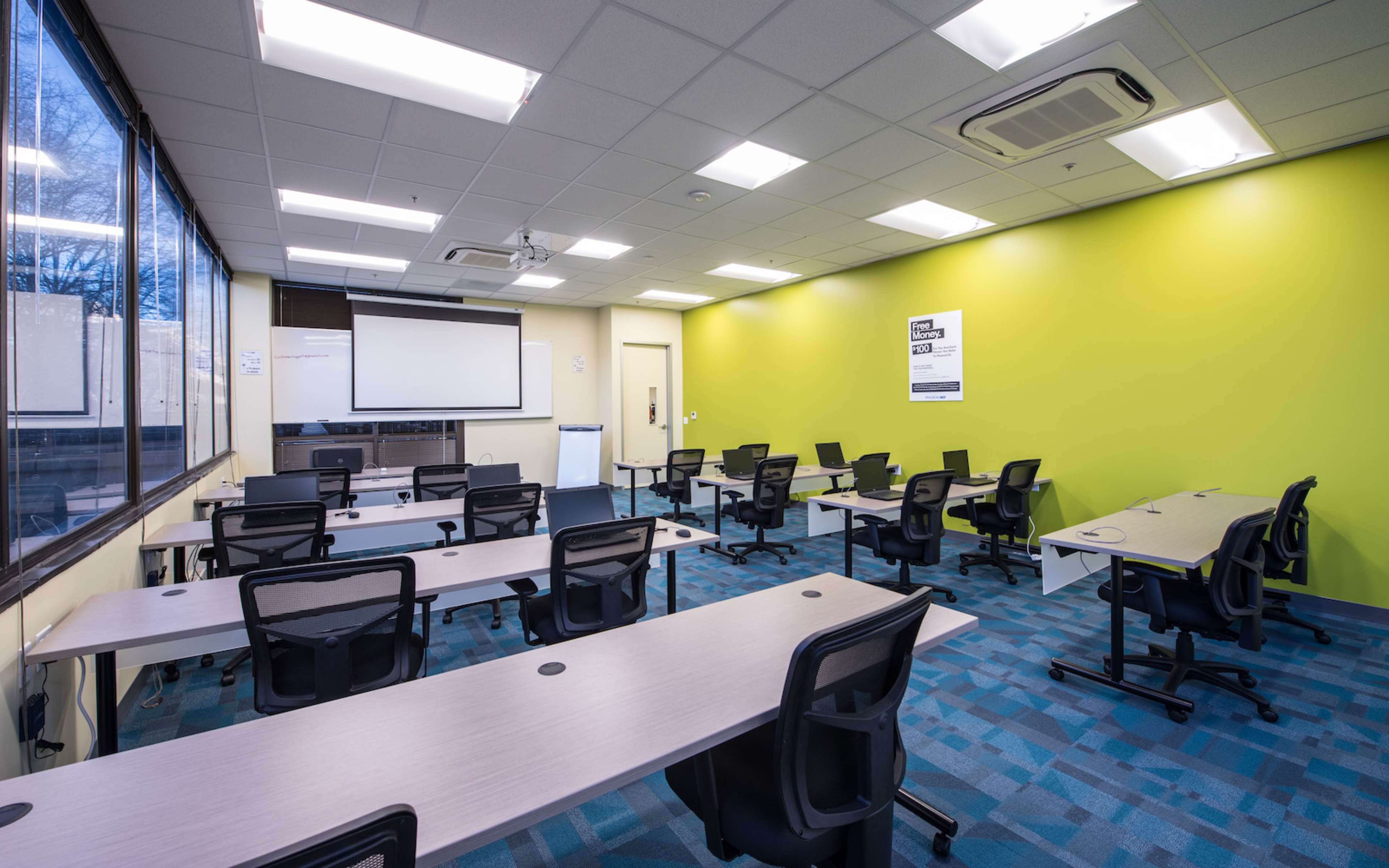 The image shows a bright classroom with several rows of desks and chairs, a whiteboard, and a projector screen against a green accent wall.