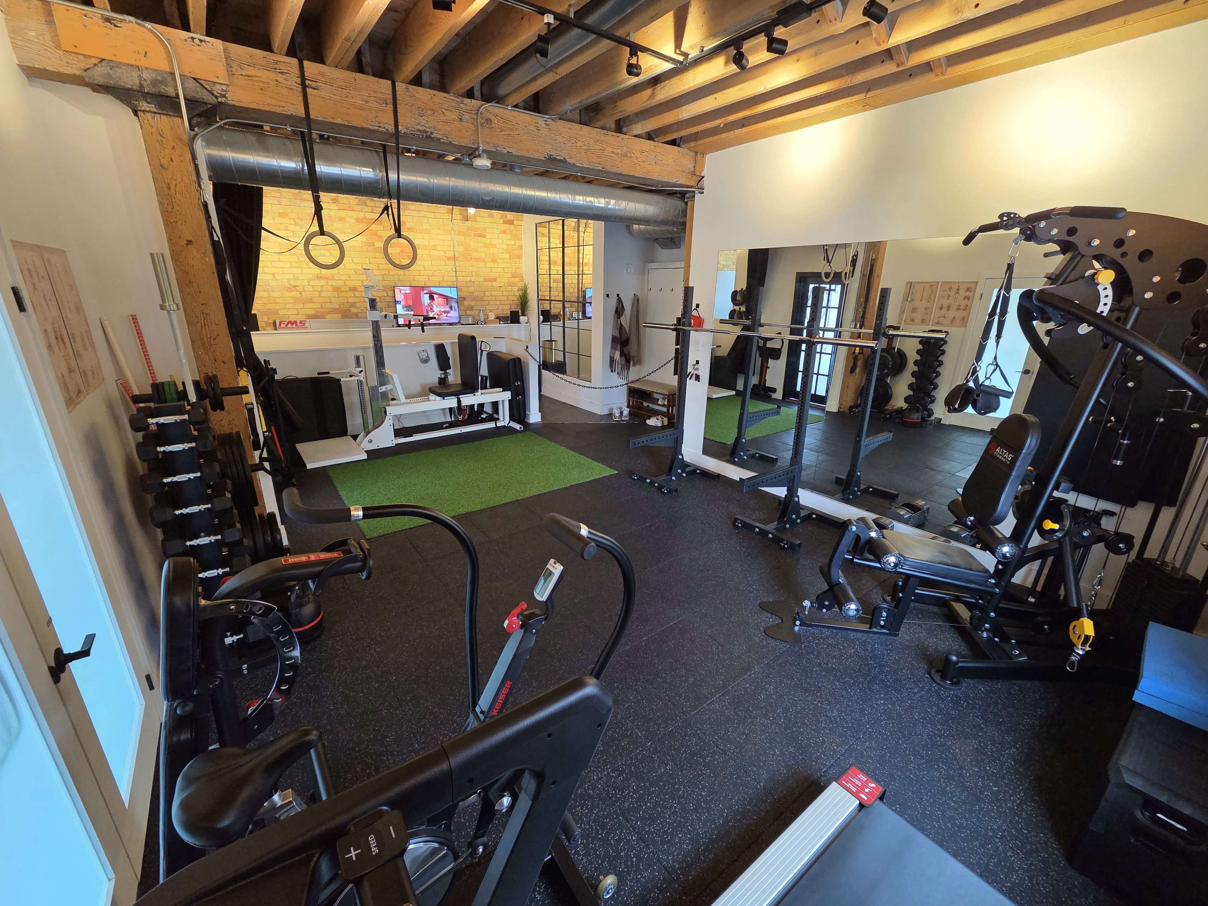 The image shows a home gym equipped with various exercise machines, weights, and exercise mats under wooden beams and bright lighting.
