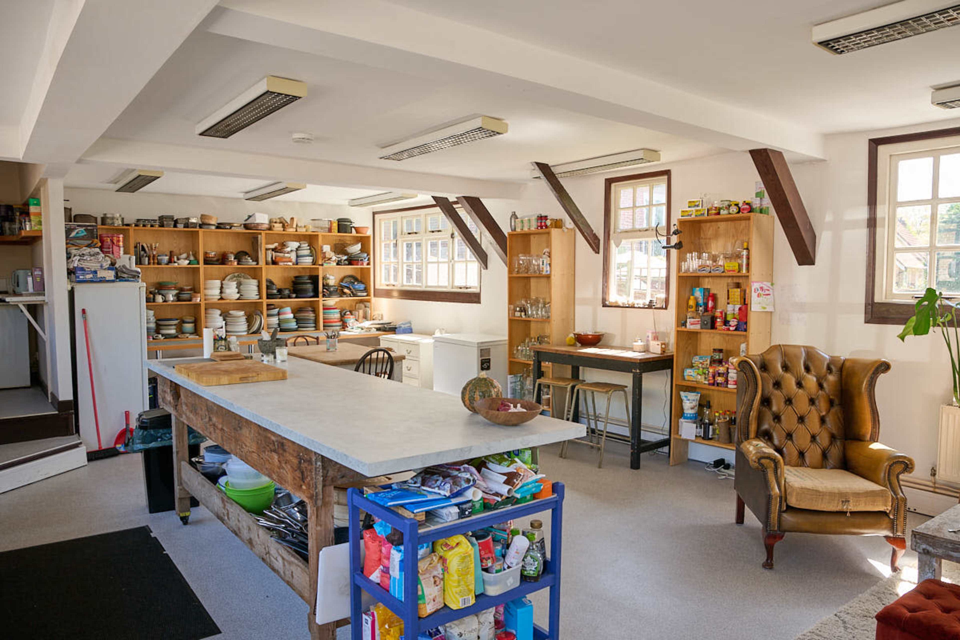 The image shows a spacious kitchen with wooden shelves filled with dishes and food supplies, an island countertop in the center, and a comfortable armchair in one corner.