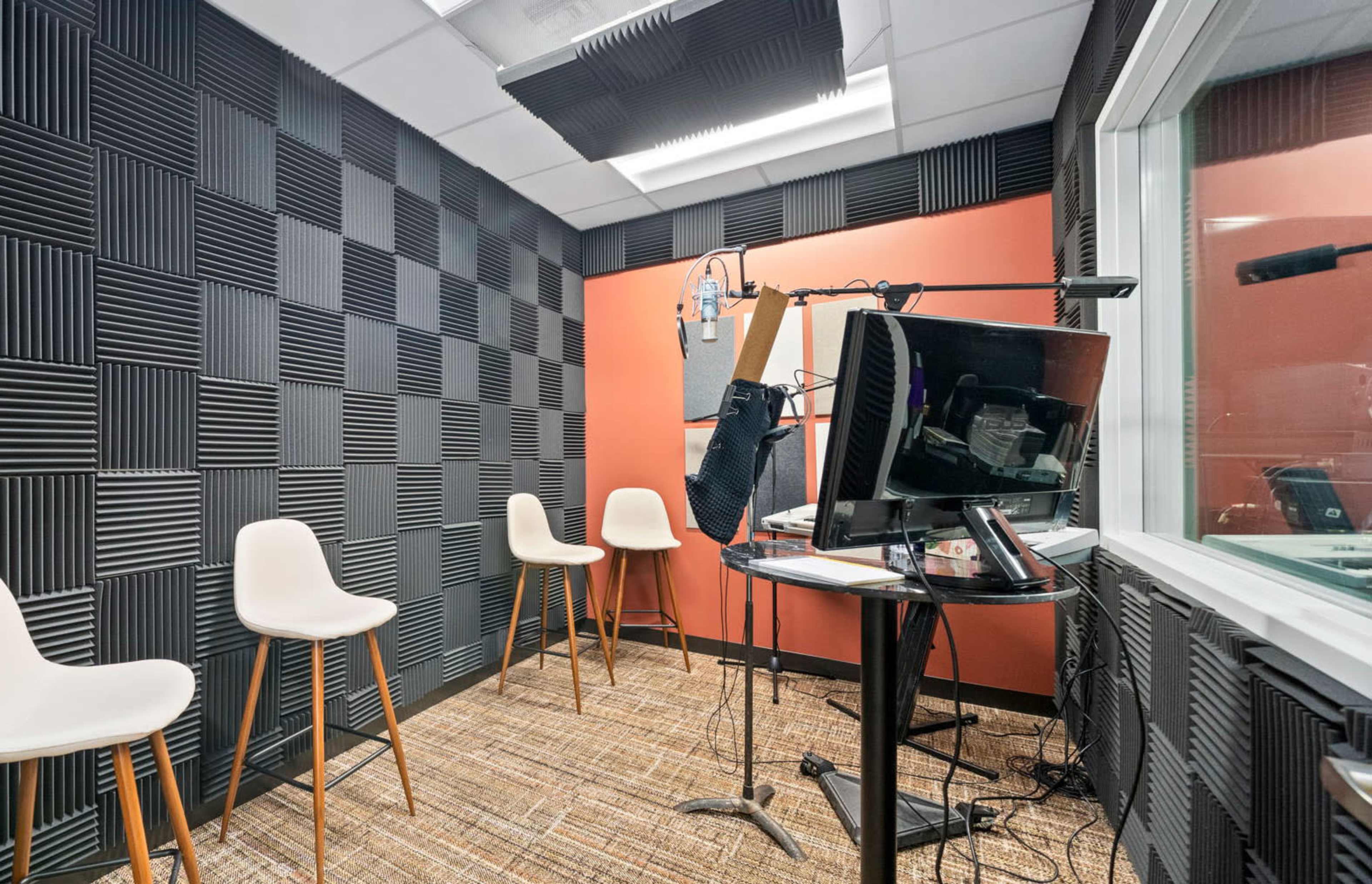 The image shows a recording studio with soundproof black acoustic panels on the walls, four white chairs, a microphone on a stand, and a monitor on a small table.