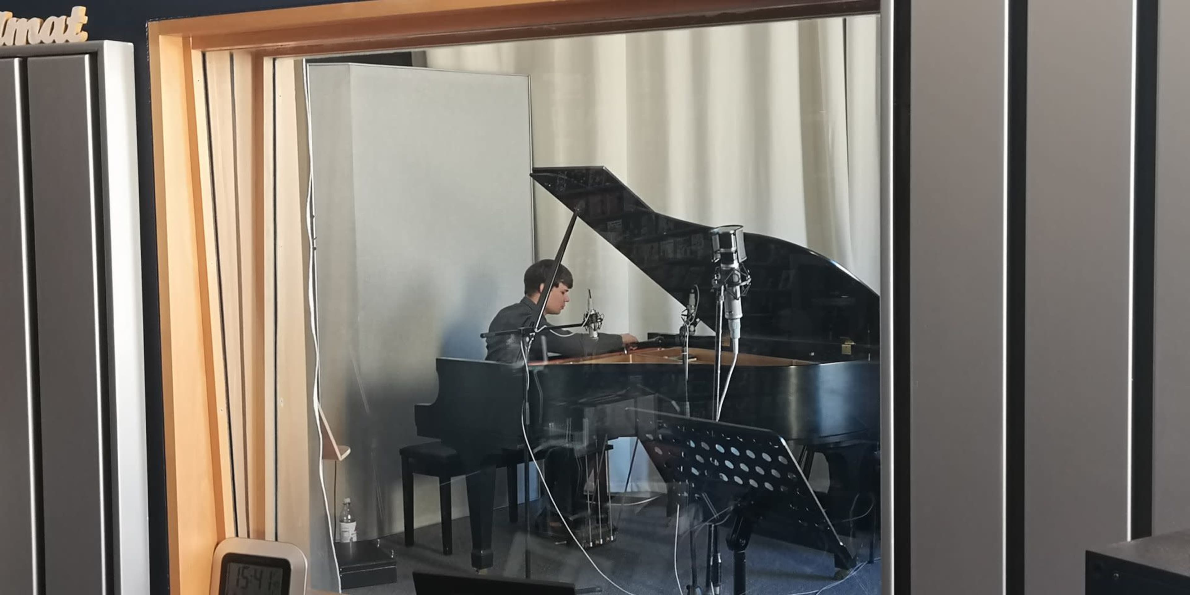 A musician is seated at a grand piano inside a recording studio, with microphones set up around them.