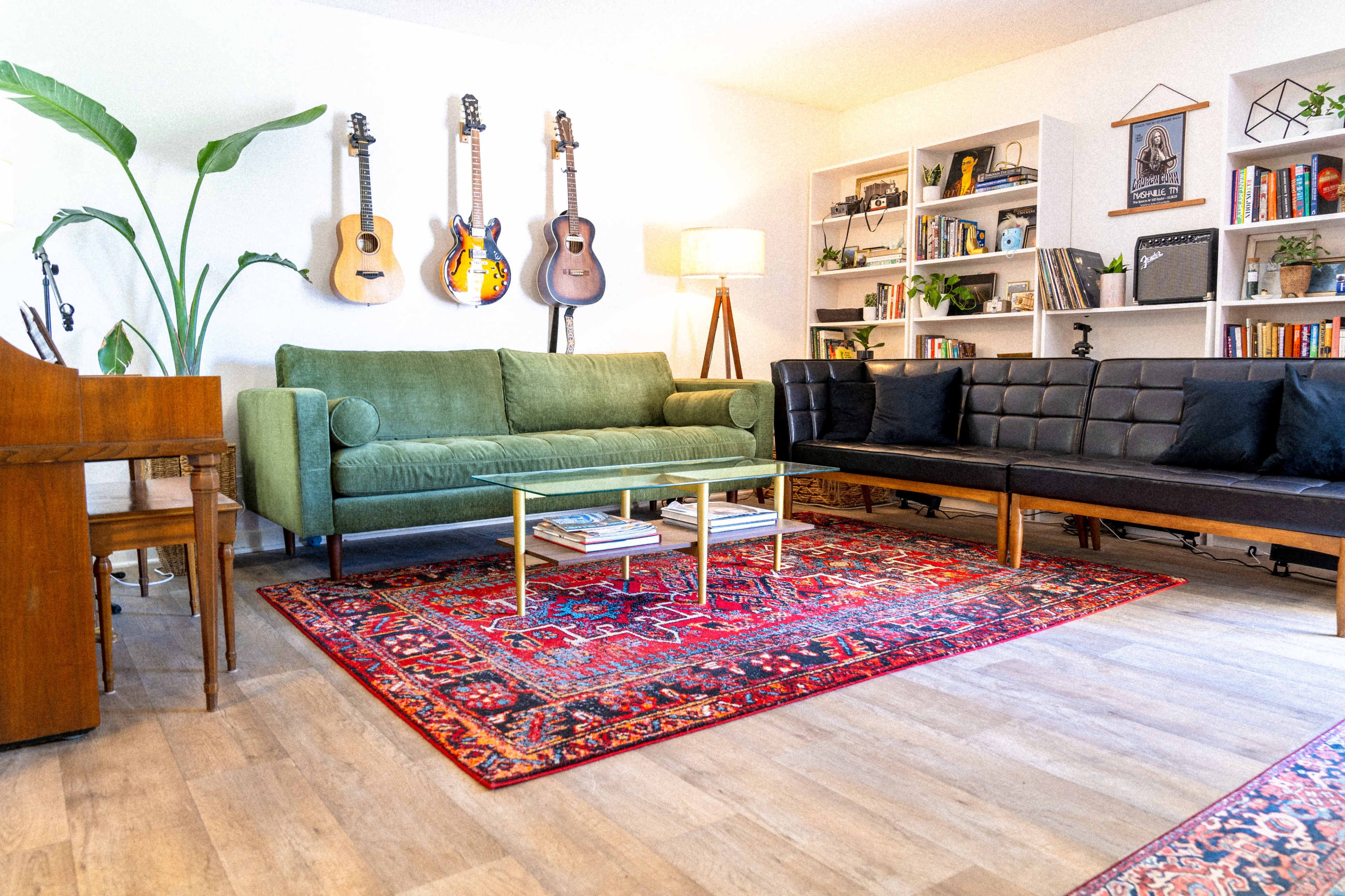 A living room features a green sofa, a black leather couch, a glass coffee table, and shelves filled with books, decorated with guitars and plants.