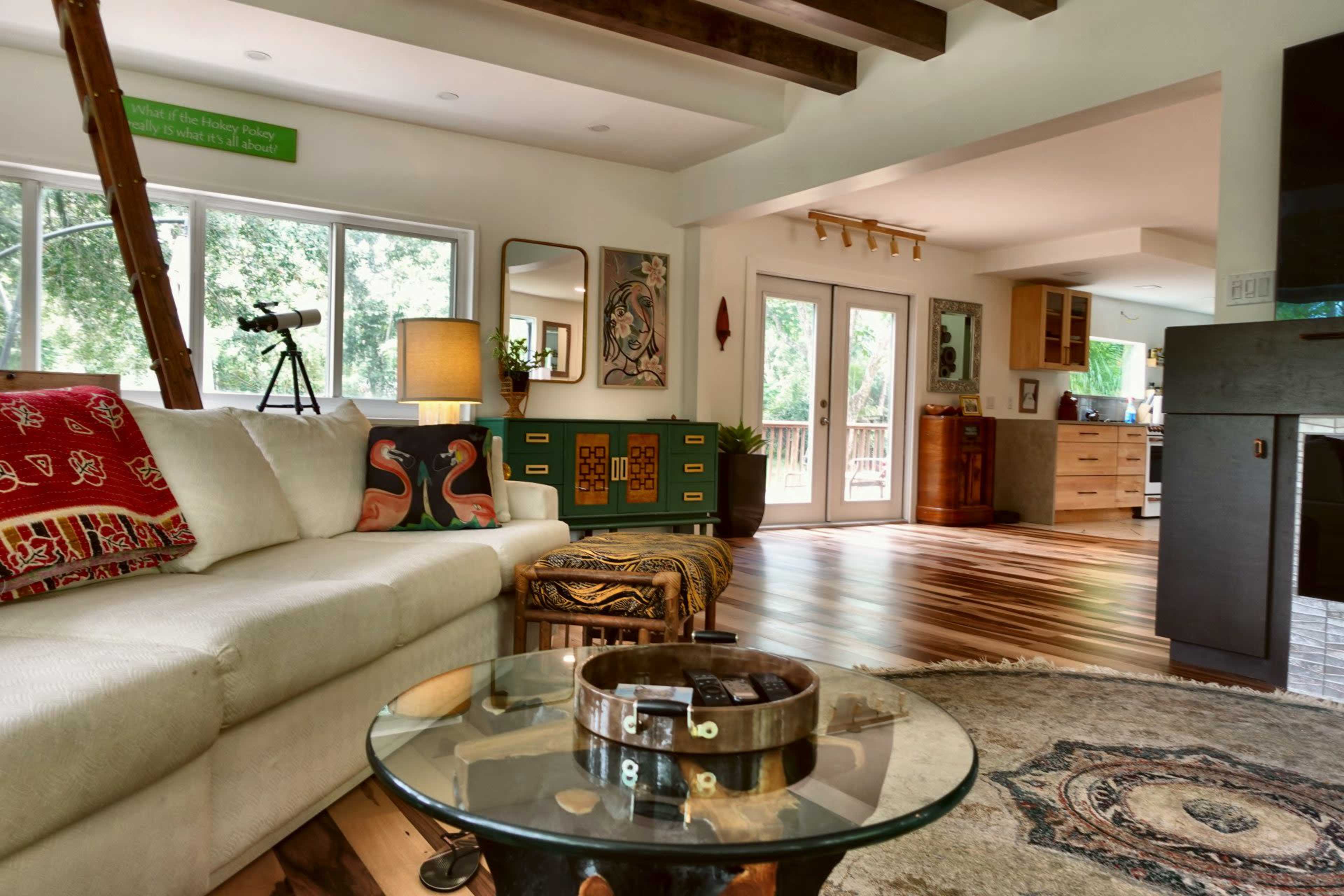 The image shows a cozy living room with a white sofa, a glass coffee table, and a view into a kitchen area featuring wooden cabinets and a large window.