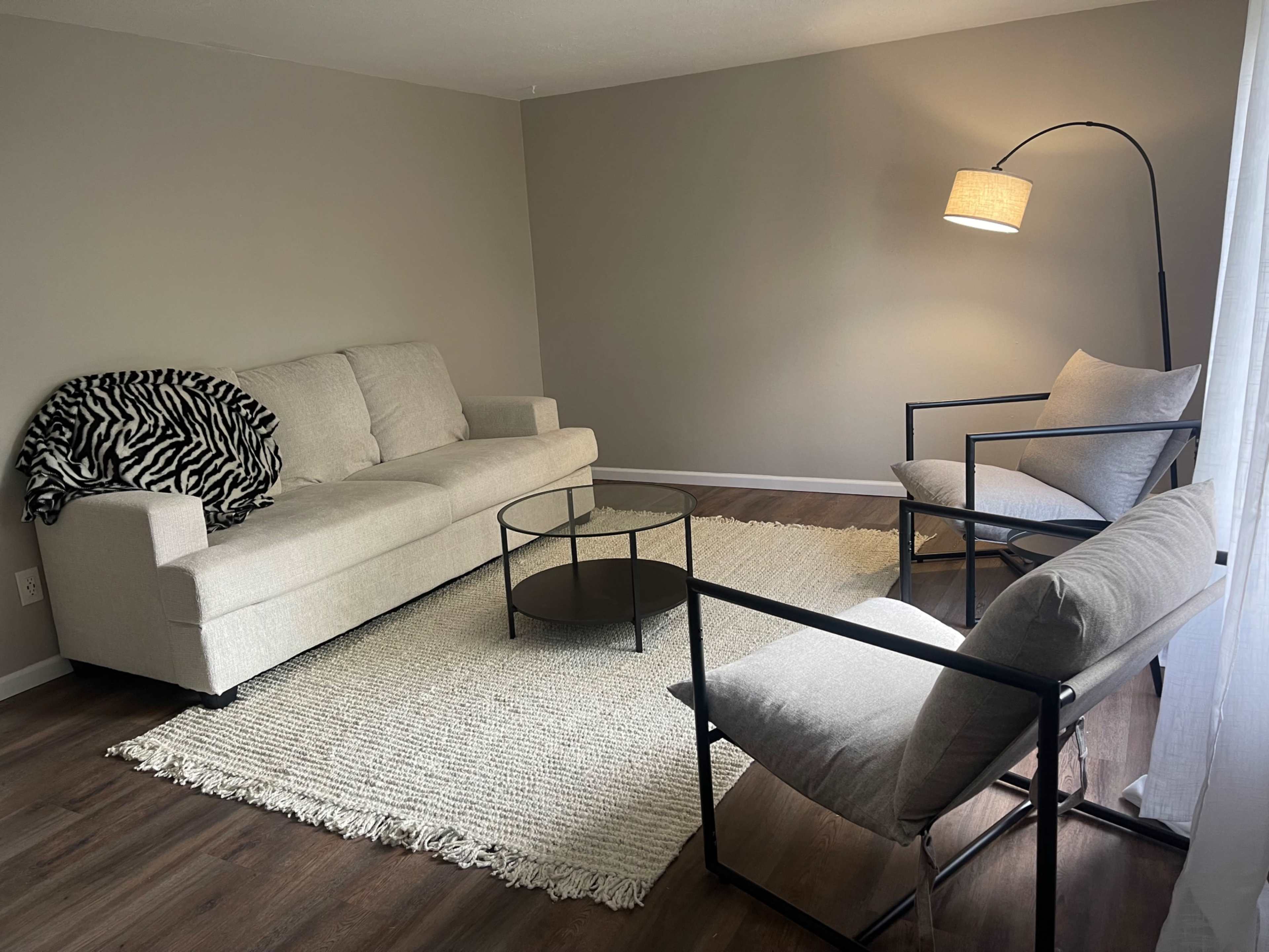 A light-colored living room featuring a sofa, two armchairs, a round coffee table, and a textured area rug.