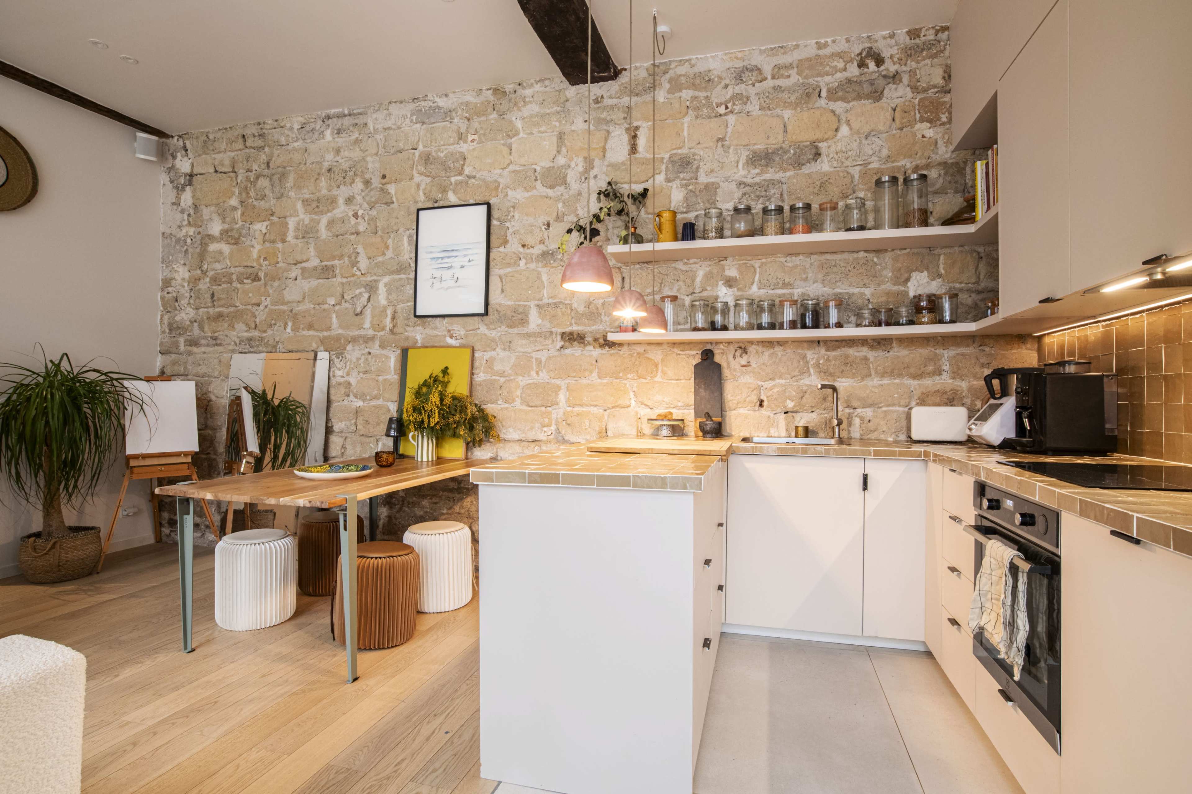 The kitchen features a rustic stone wall, modern cabinetry, and a wooden dining table set against a backdrop of decorative plants and various kitchen items.