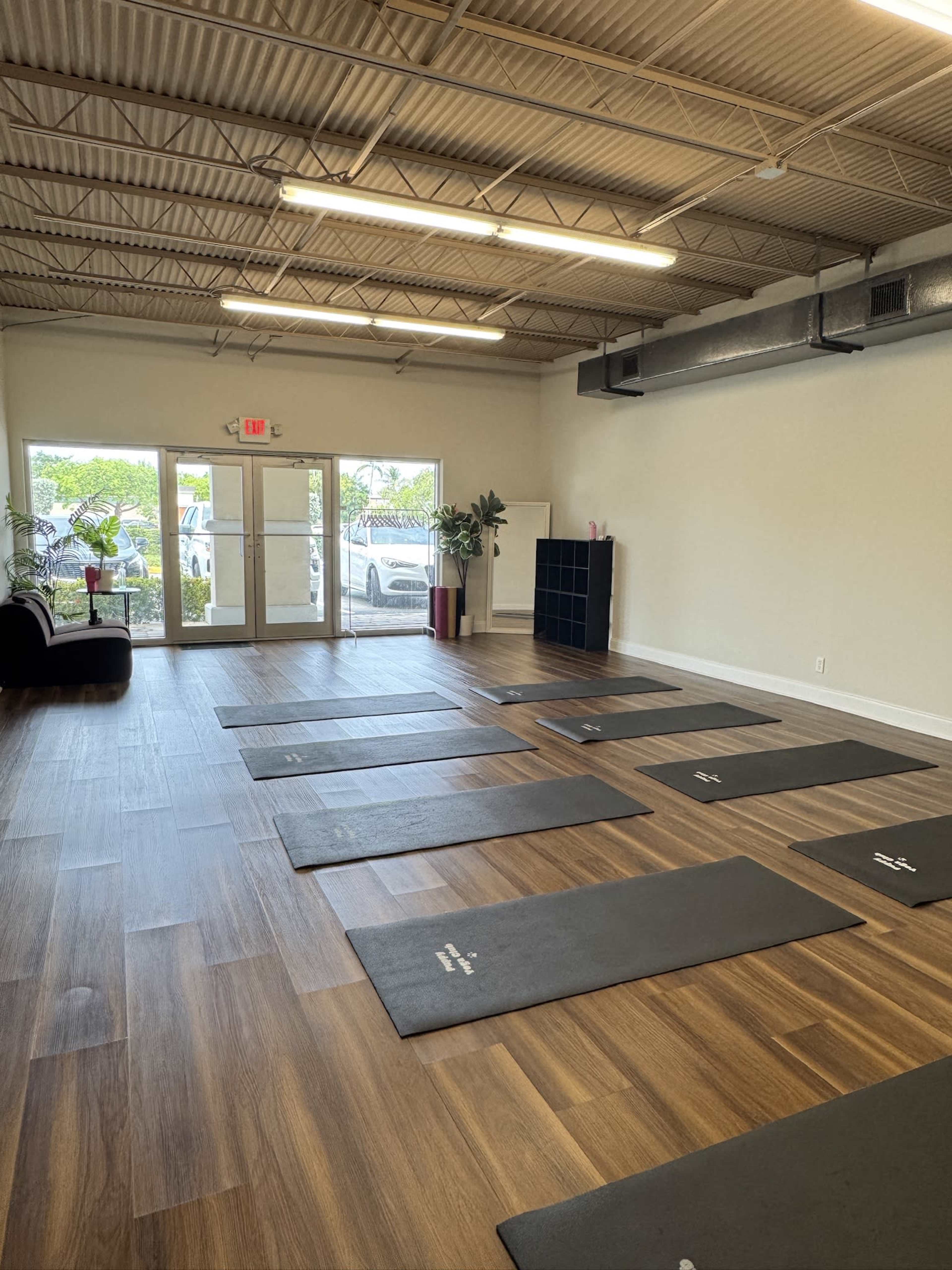 The image shows a spacious room with yoga mats arranged on a wooden floor, large windows letting in natural light, and a few potted plants in the corners.