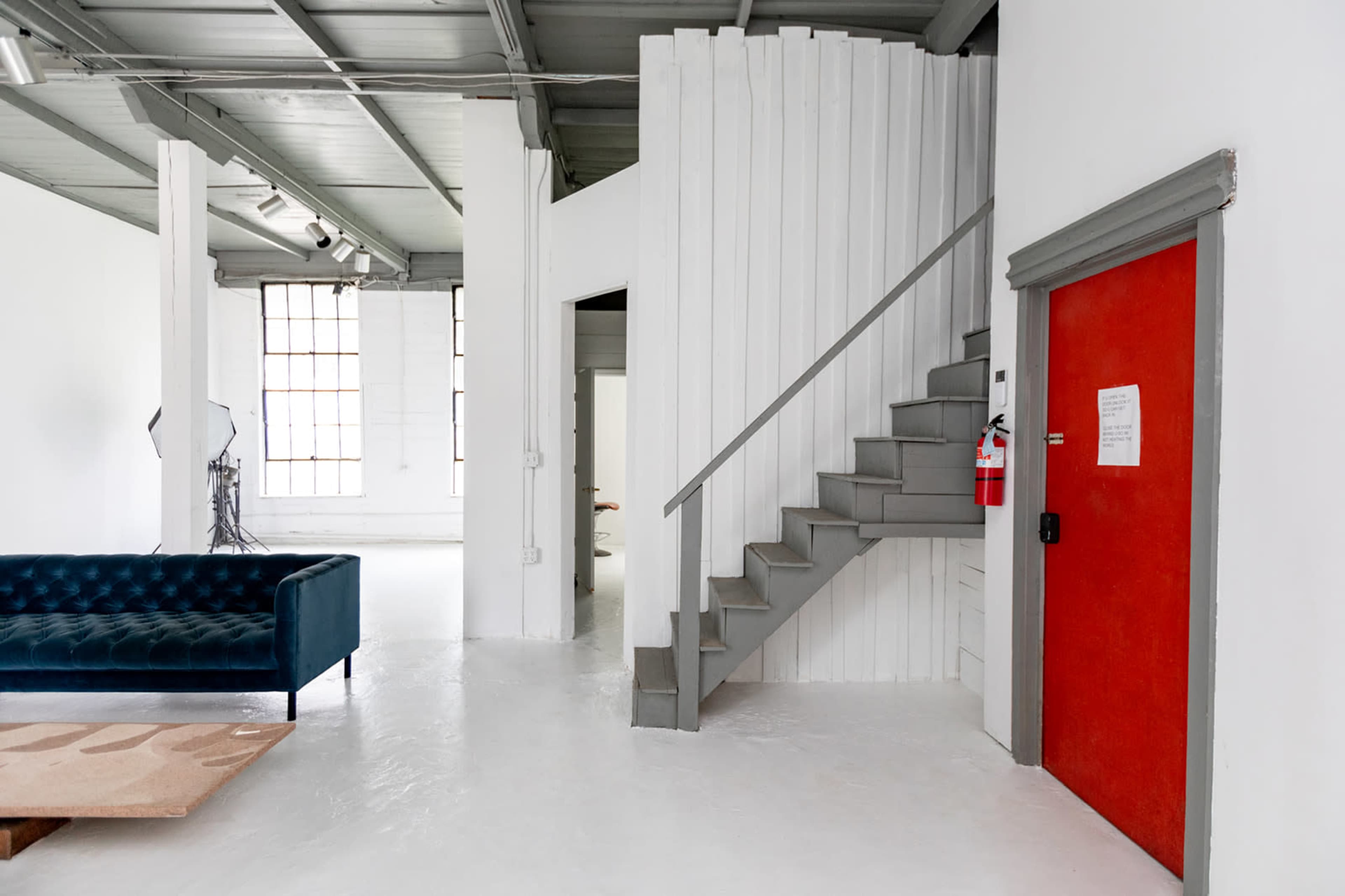The image shows a bright, minimalist interior featuring a blue velvet couch, a staircase leading to an upper level, and a red door with a notice on it.