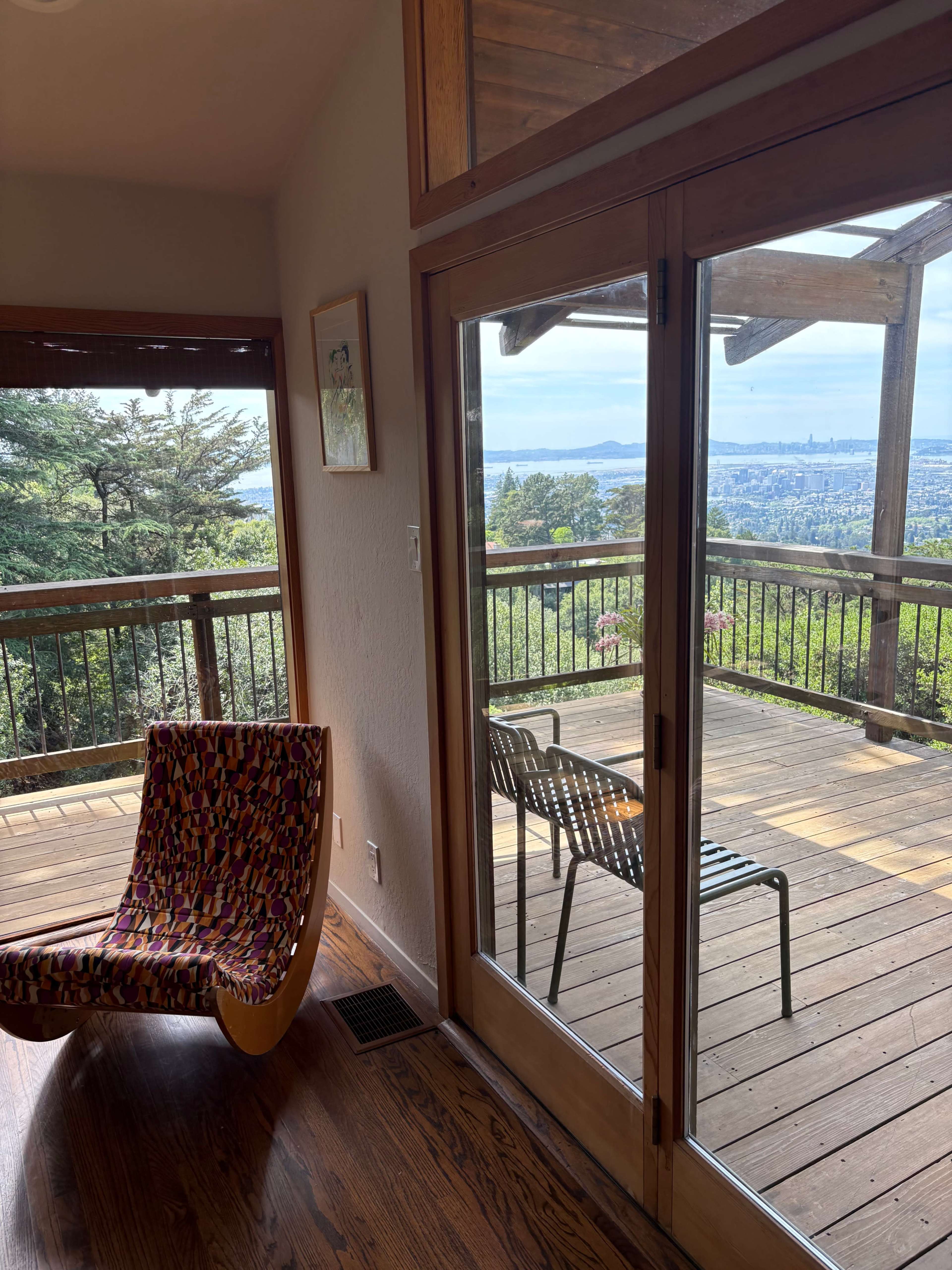 The image shows a spacious room with a colorful chair and large sliding glass doors that open to a deck overlooking a view of a city and mountains.