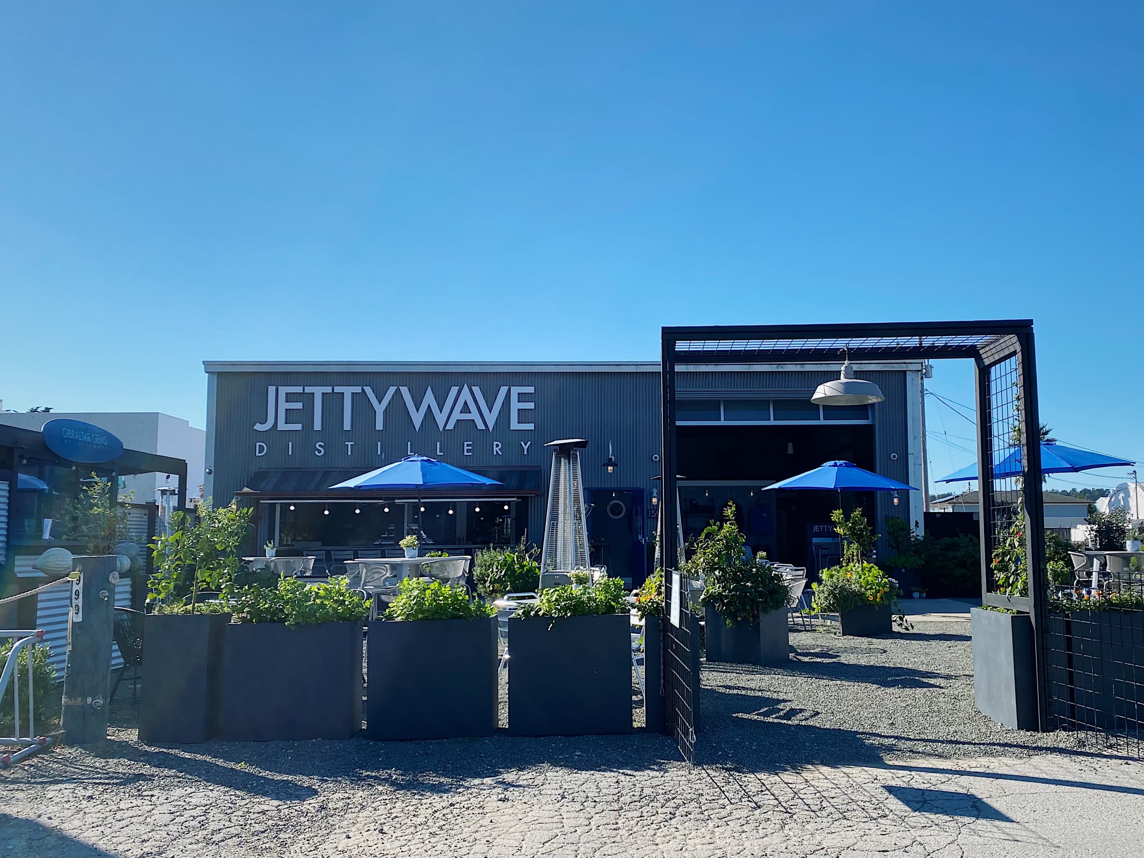 The exterior of Jetty Wave Distillery, featuring a modern building with blue umbrellas and planters in a sunny outdoor space.