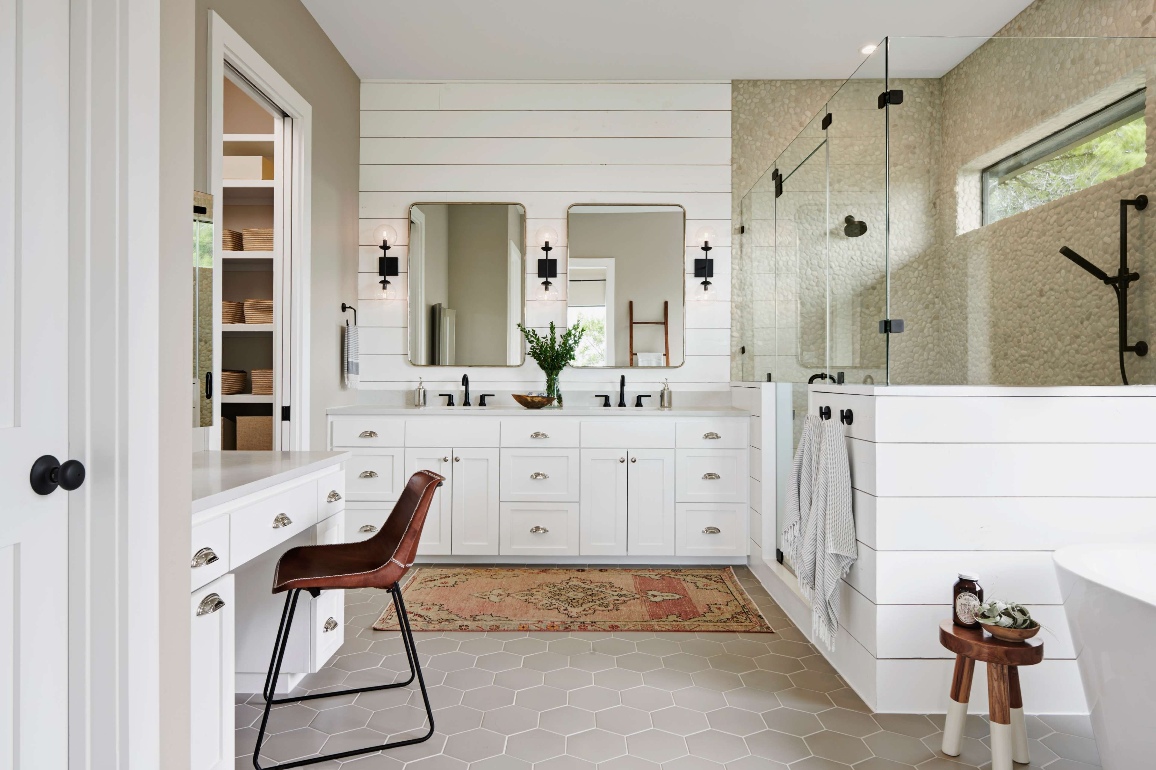 A modern bathroom with white cabinetry, dual mirrors, and a glass shower, complemented by a round side table and an intricate rug on the floor.