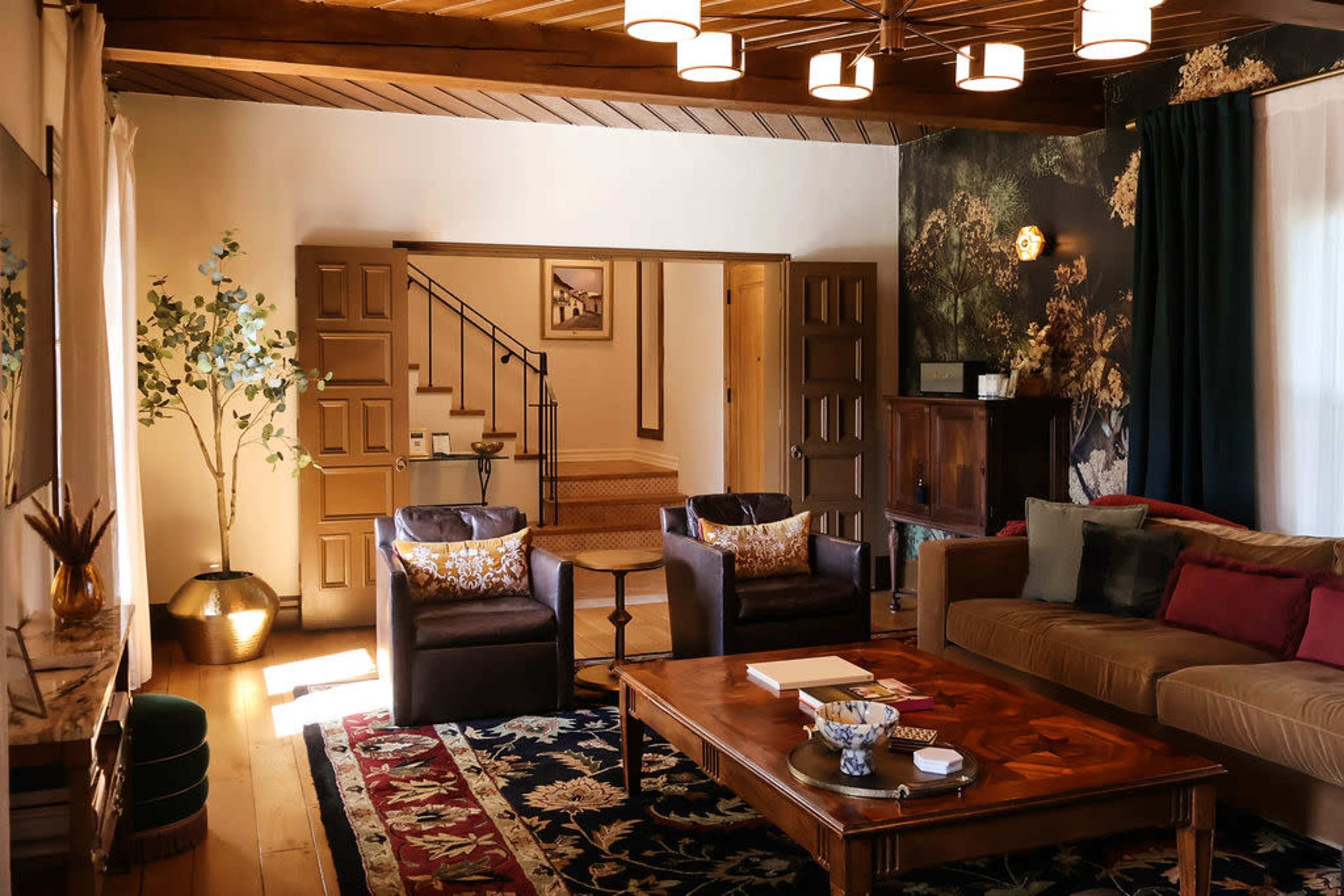 The image shows a cozy living room featuring two armchairs, a large coffee table, and a staircase leading to another area, with decorative plants and a patterned rug.