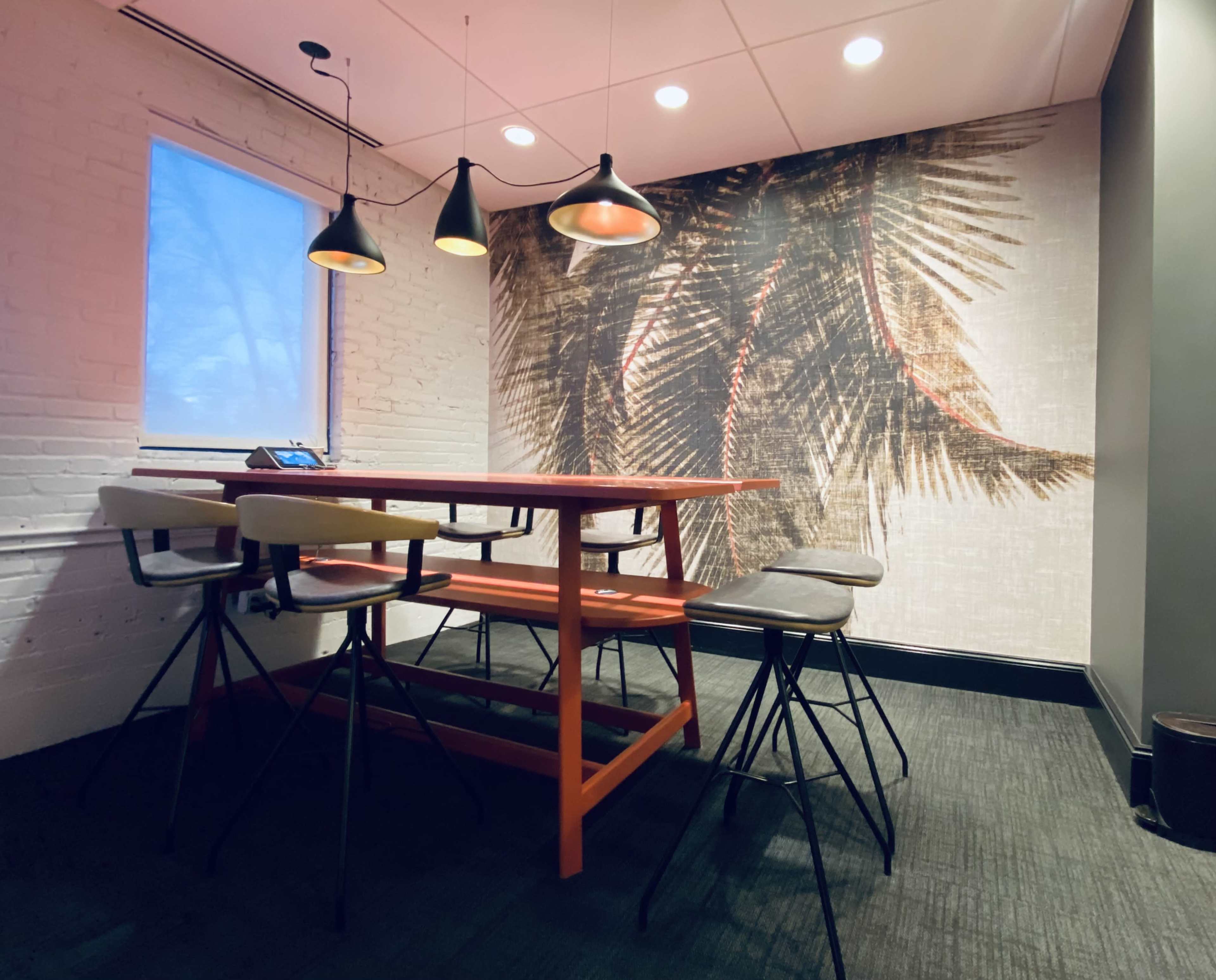 The image shows a modern meeting room featuring a wooden table surrounded by different colored chairs and a decorative wall with a large tropical plant mural.