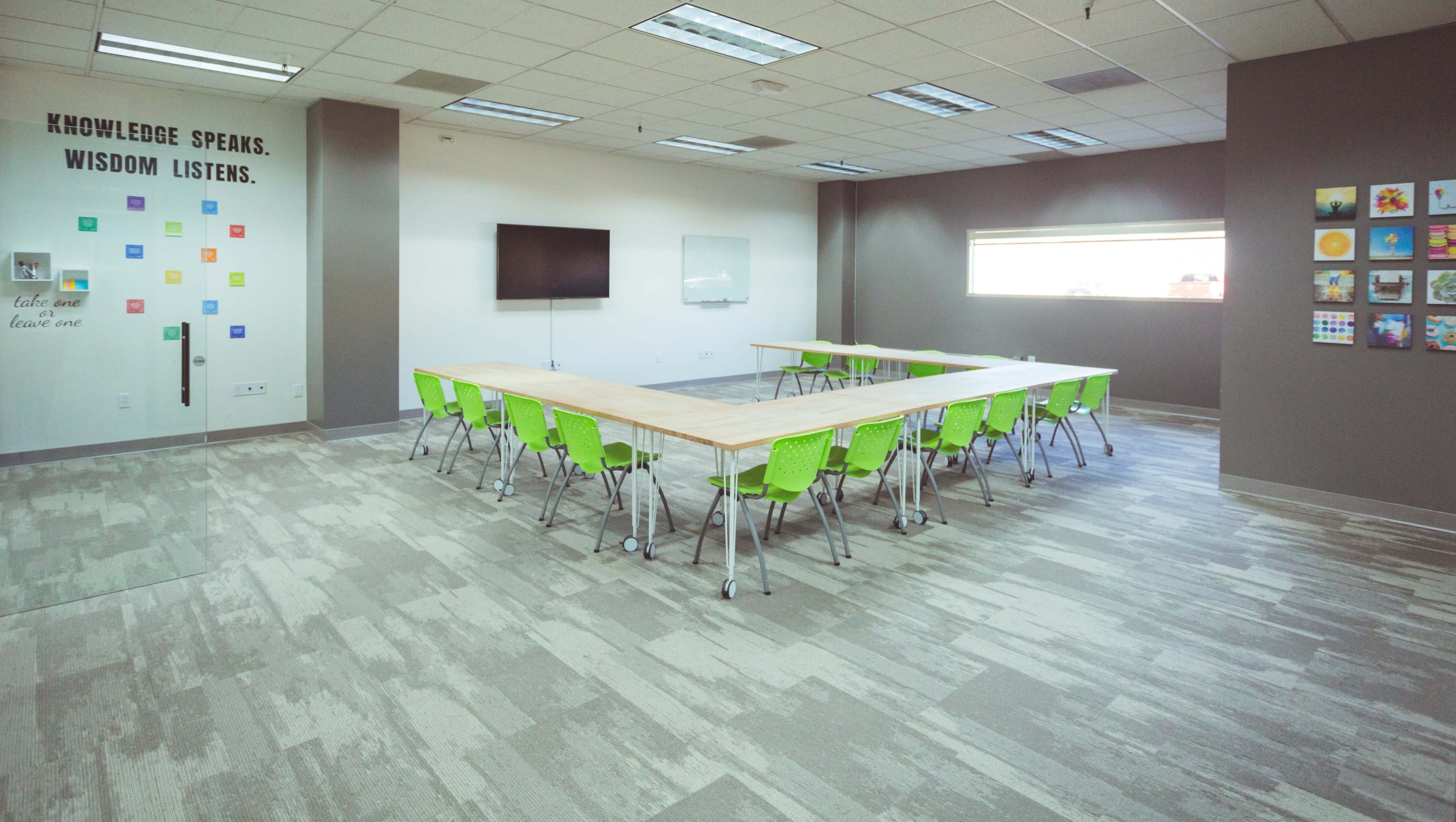 A modern conference room features a large wooden table surrounded by green chairs, with a wall displaying colorful artwork and a screen for presentations.