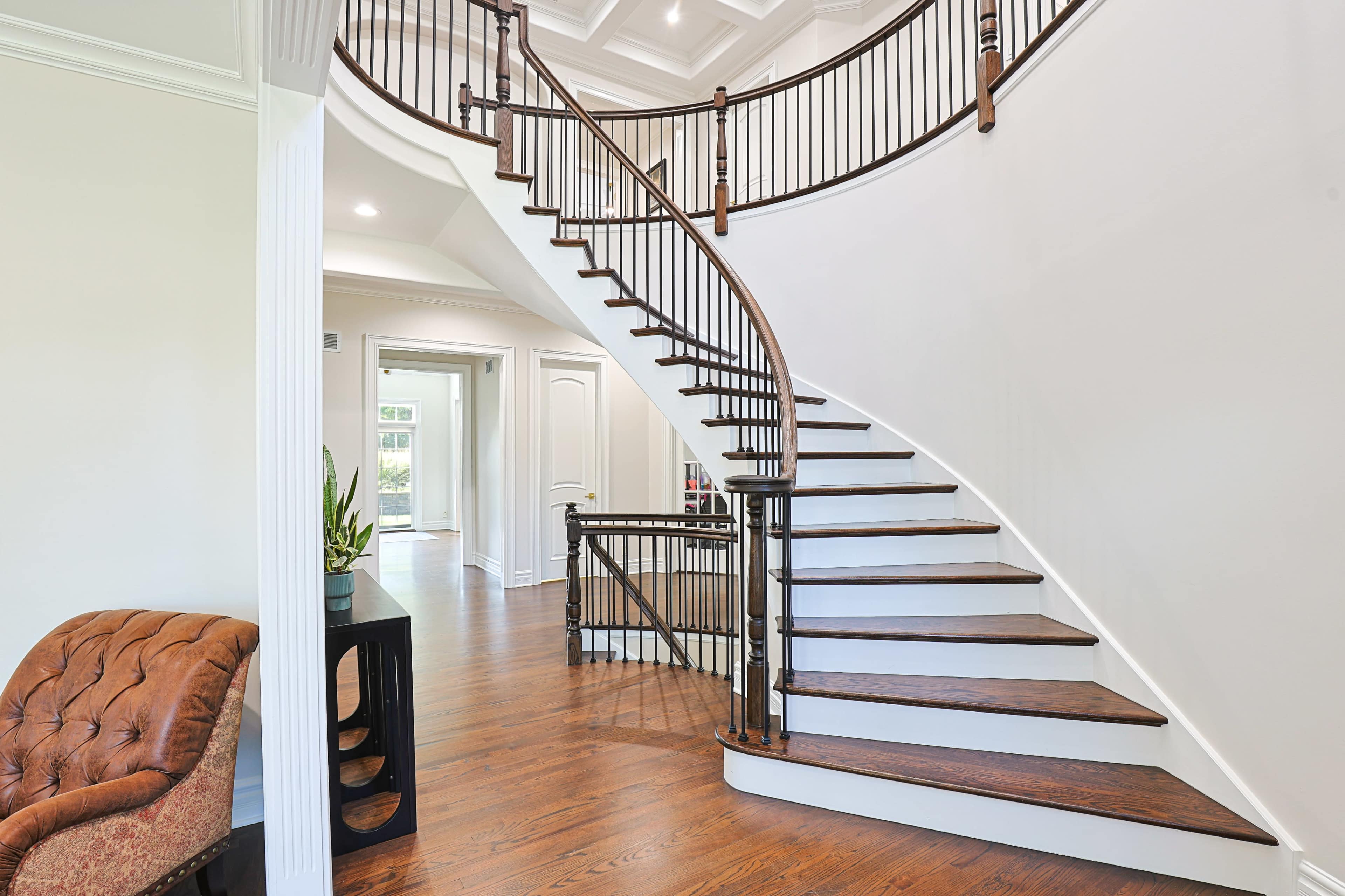 The image shows a curved wooden staircase with a black railing in a well-lit foyer, featuring a tufted brown armchair and a small table nearby.