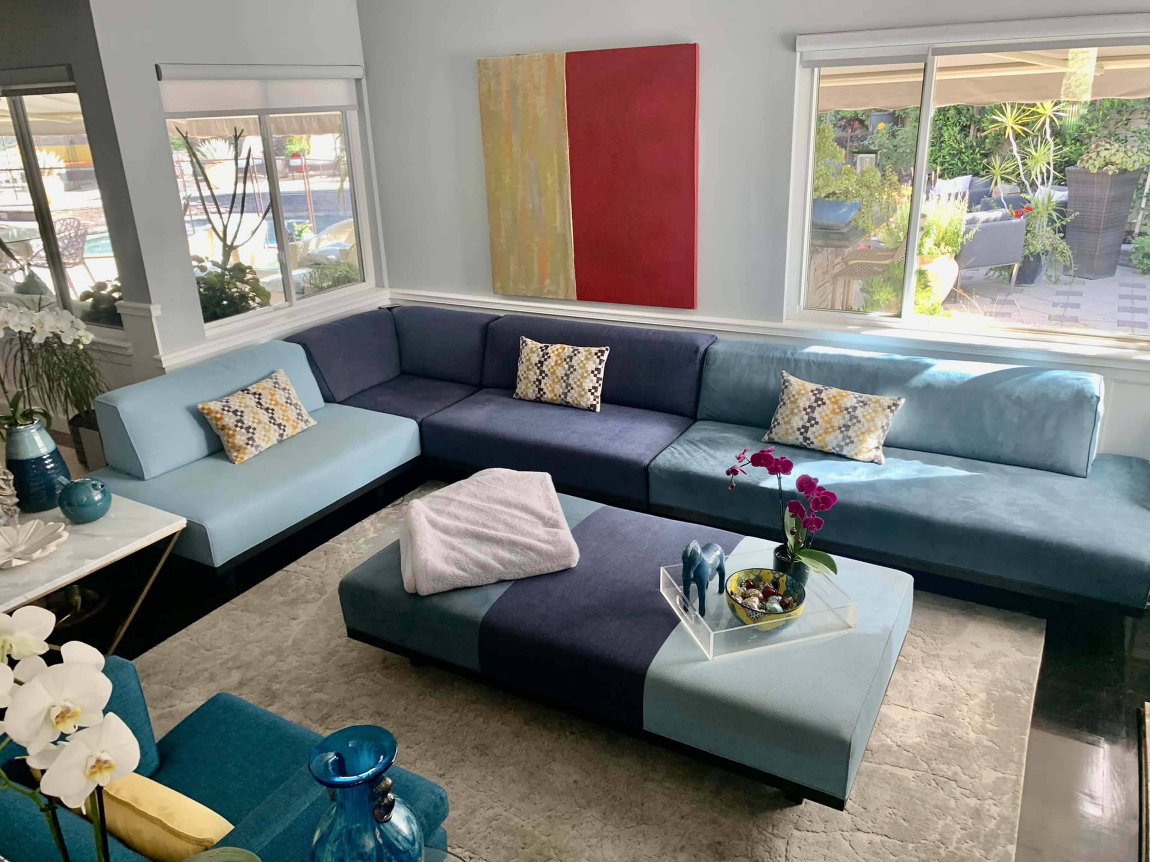A spacious living room features a large blue sectional sofa with decorative pillows, a central coffee table, and large windows with views of an outdoor space.