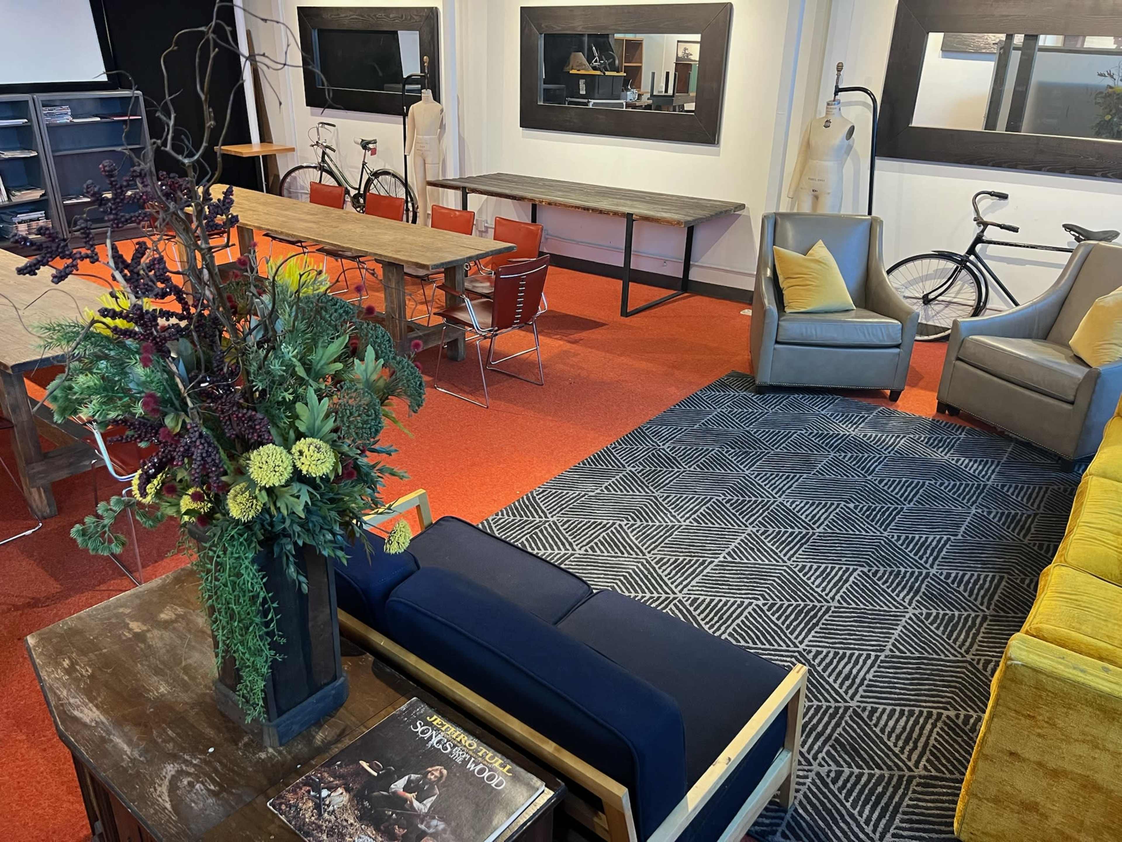 The image shows a modern, open workspace featuring a mix of seating areas, tables, and decorative plants, with bicycles and mannequins positioned in the background.