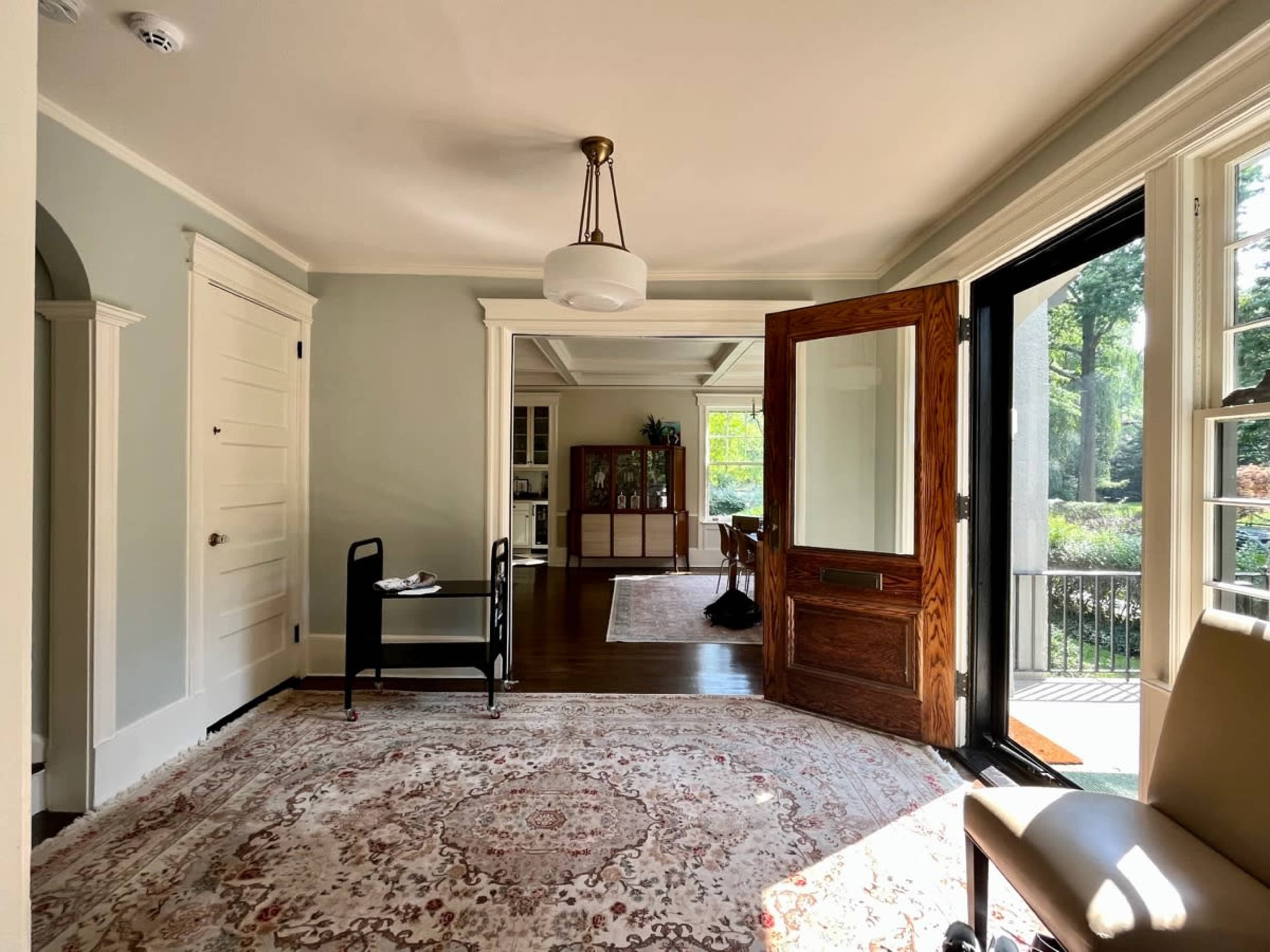 A well-lit interior room featuring a large open door leading to an outdoor space, a small black cart, and wooden furniture visible in the adjacent room.