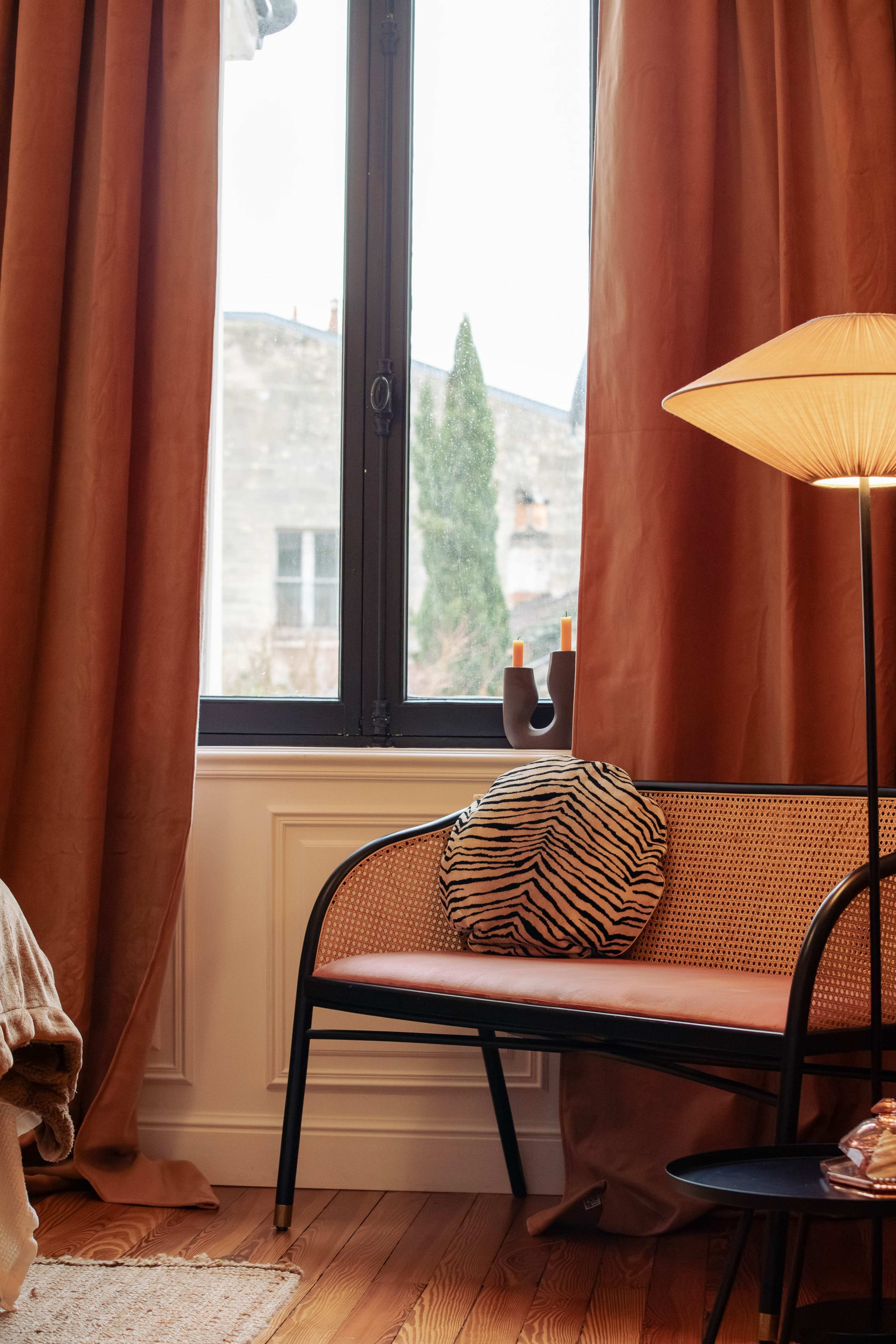 A cozy interior scene features a bench with a striped cushion beside a tall lamp, draped with long, brown curtains and a view of the outside through a large window.