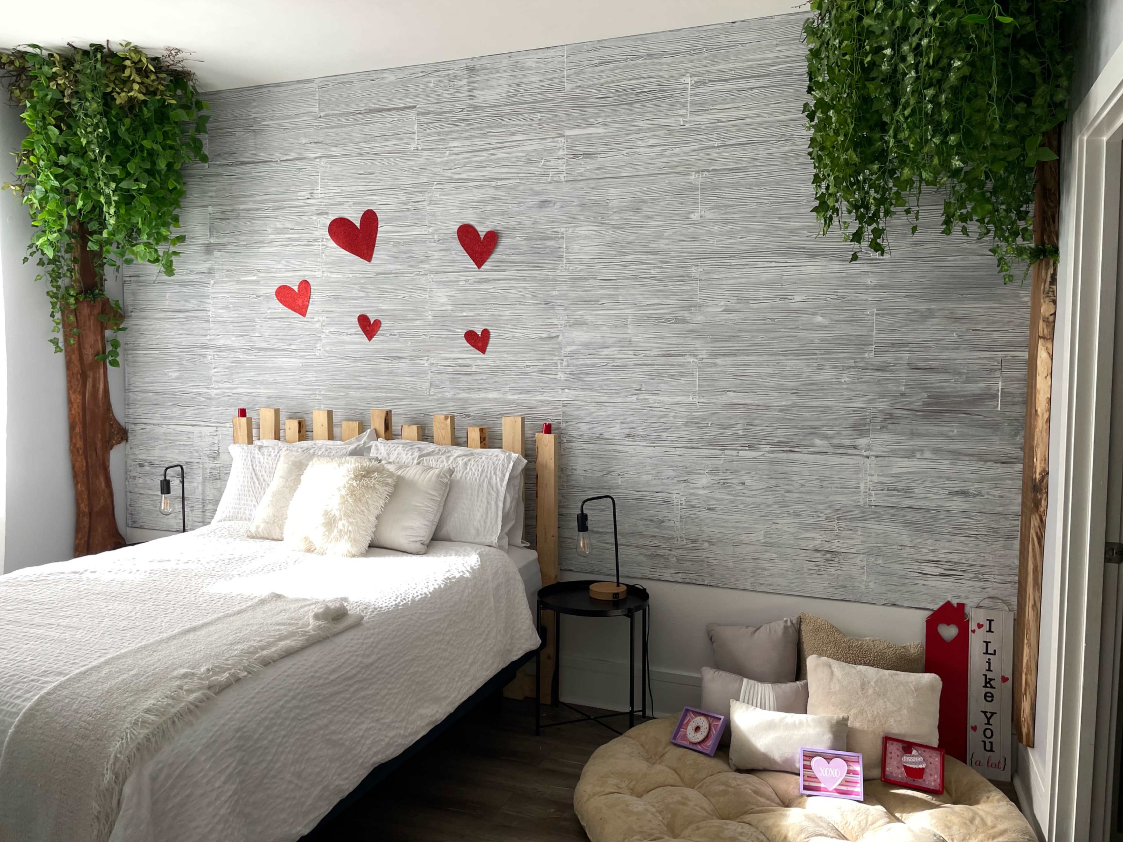 A modern bedroom featuring a wooden bed with a light-colored blanket, two decorative trees on either side of the bed, and a wall adorned with red heart cutouts.