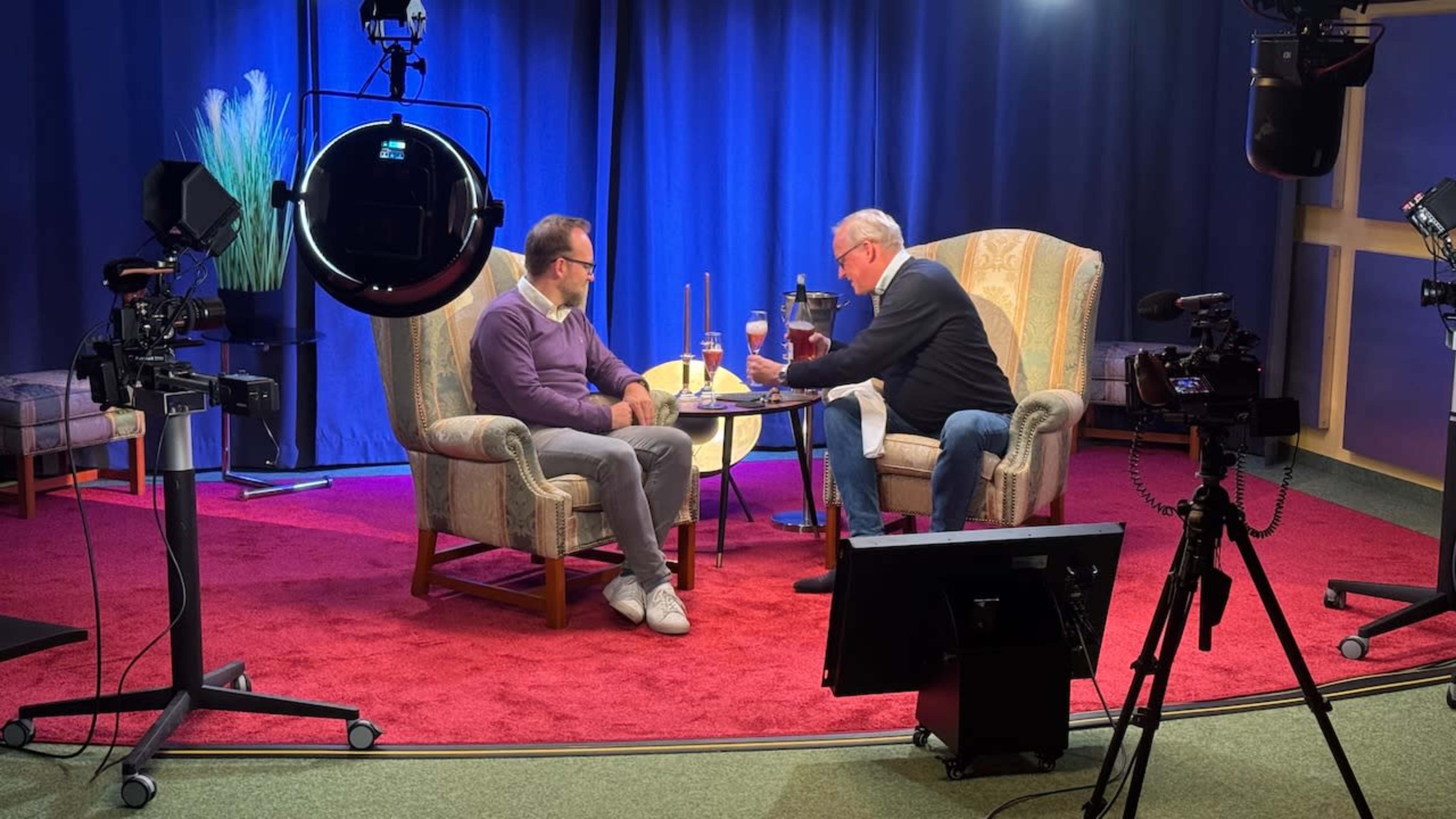 Two men sit at a small table in a television studio, discussing over drinks while surrounded by cameras and studio lighting.