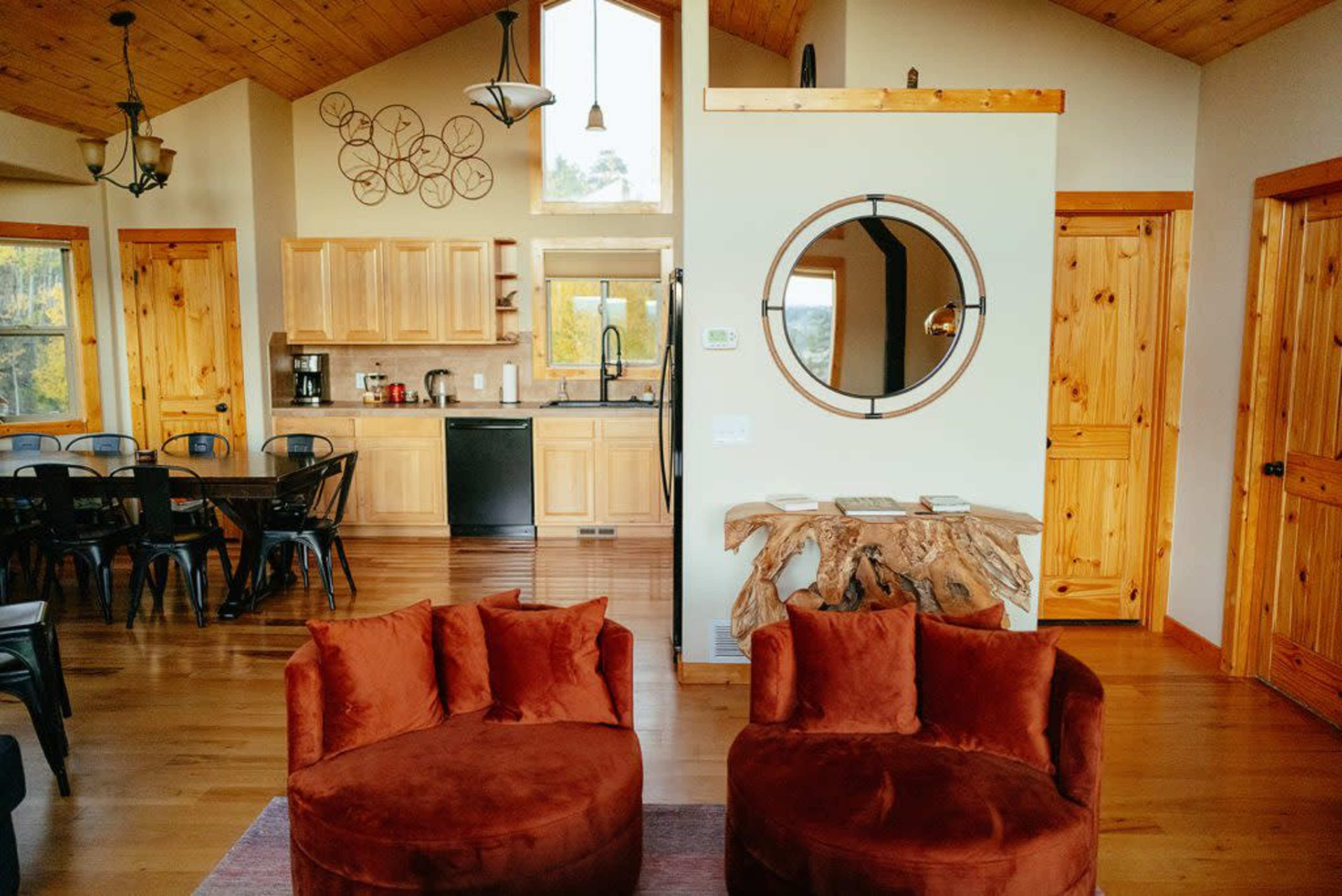 The image shows a cozy living space featuring two round, rust-colored chairs in the foreground, with a modern kitchen and dining area in the background, highlighted by wooden cabinetry and a large window.