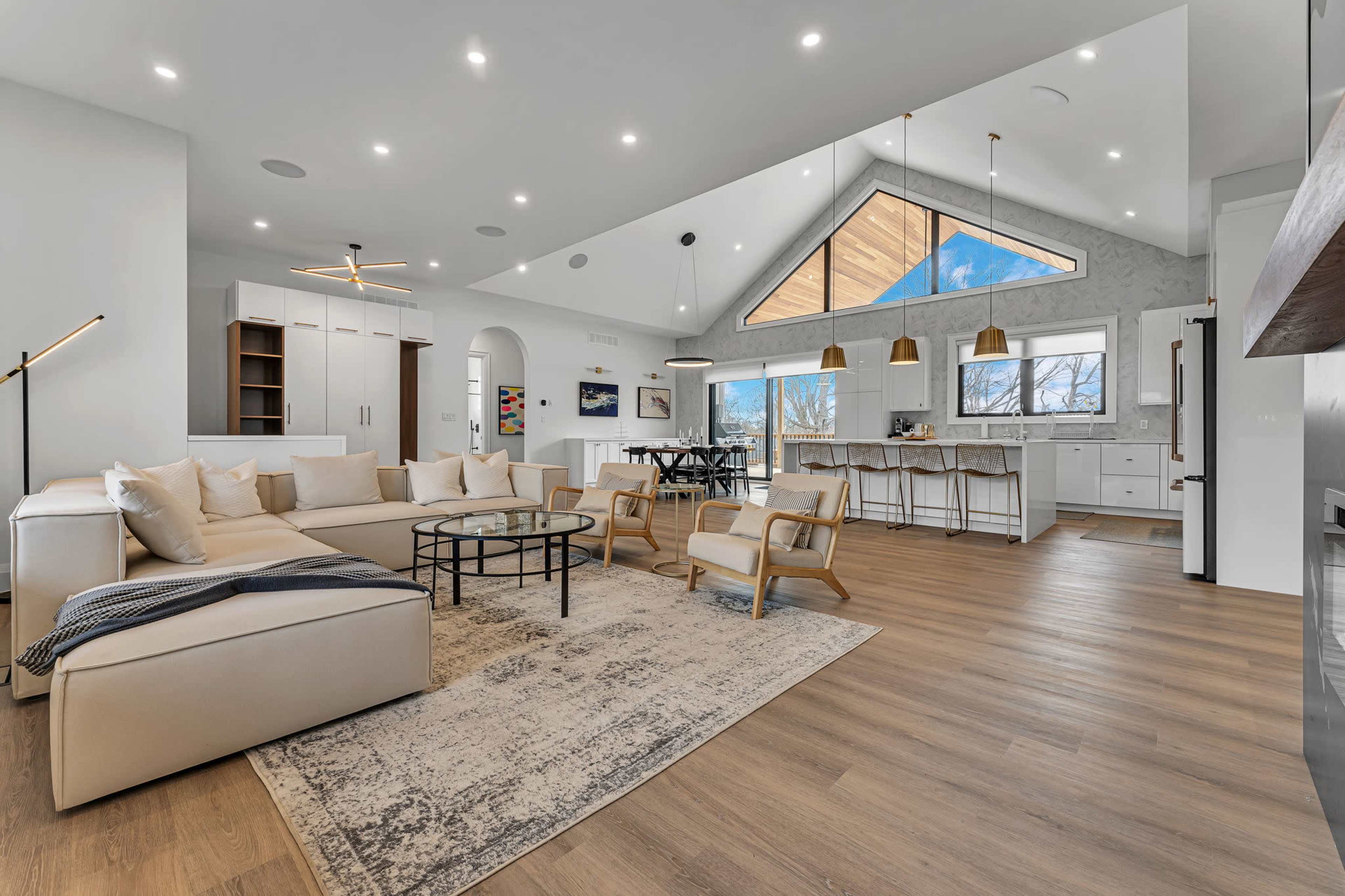 The modern living area features a sectional sofa and a circular coffee table, with an open layout leading to a dining space and kitchen, highlighted by large windows and pendant lighting.