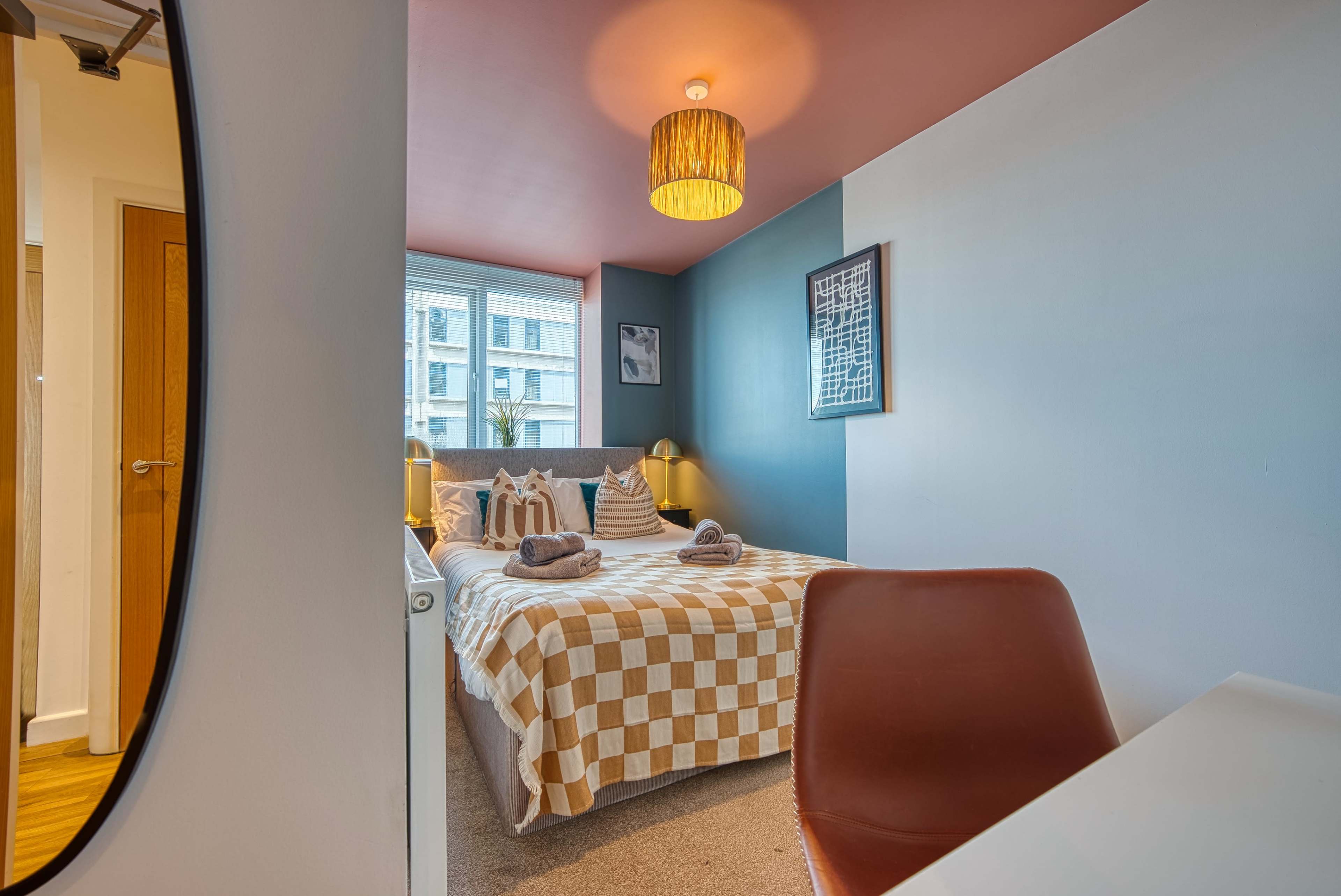 A cozy bedroom featuring a checked bedspread, a round mirror, and a desk chair, with a window allowing natural light to enter.