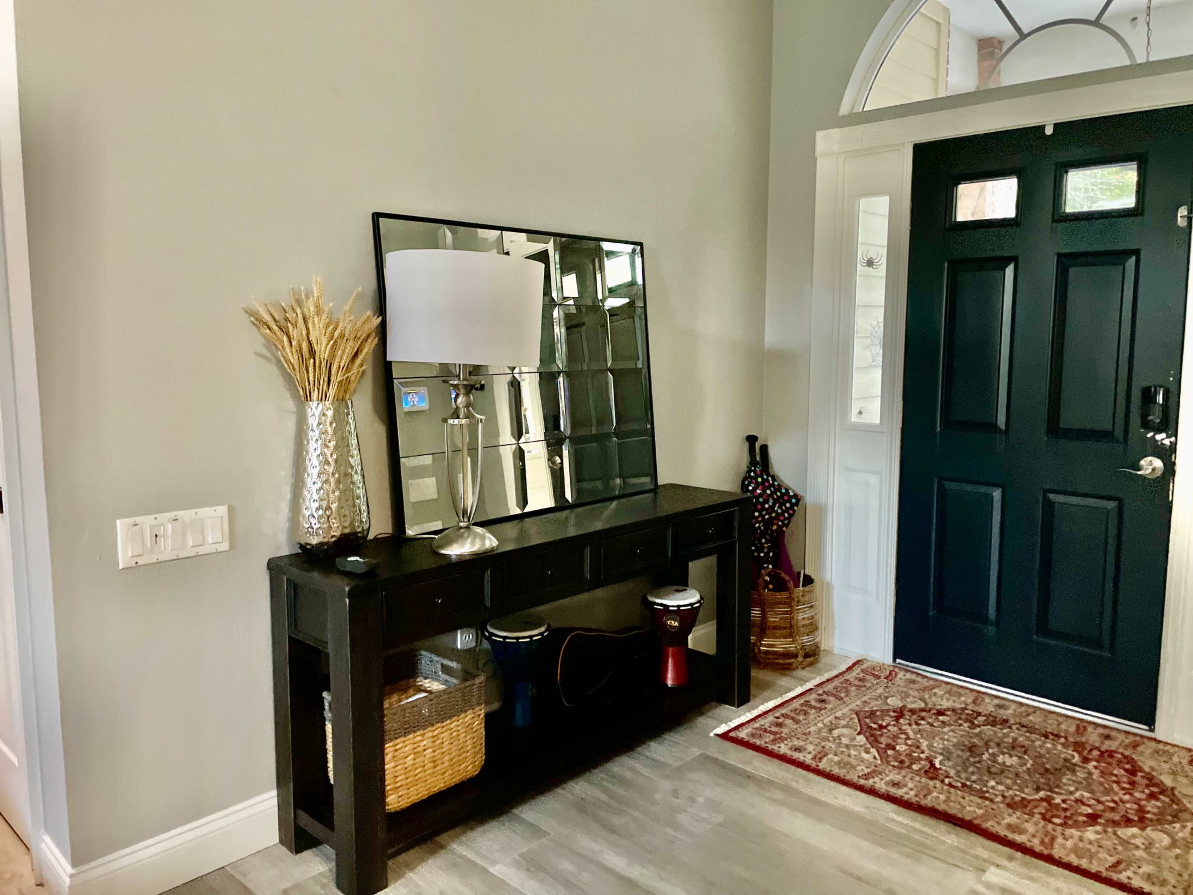 The image shows a hallway entrance featuring a dark wooden console table with a large mirror, a lamp, decorative branches, and a rug beside a black front door.