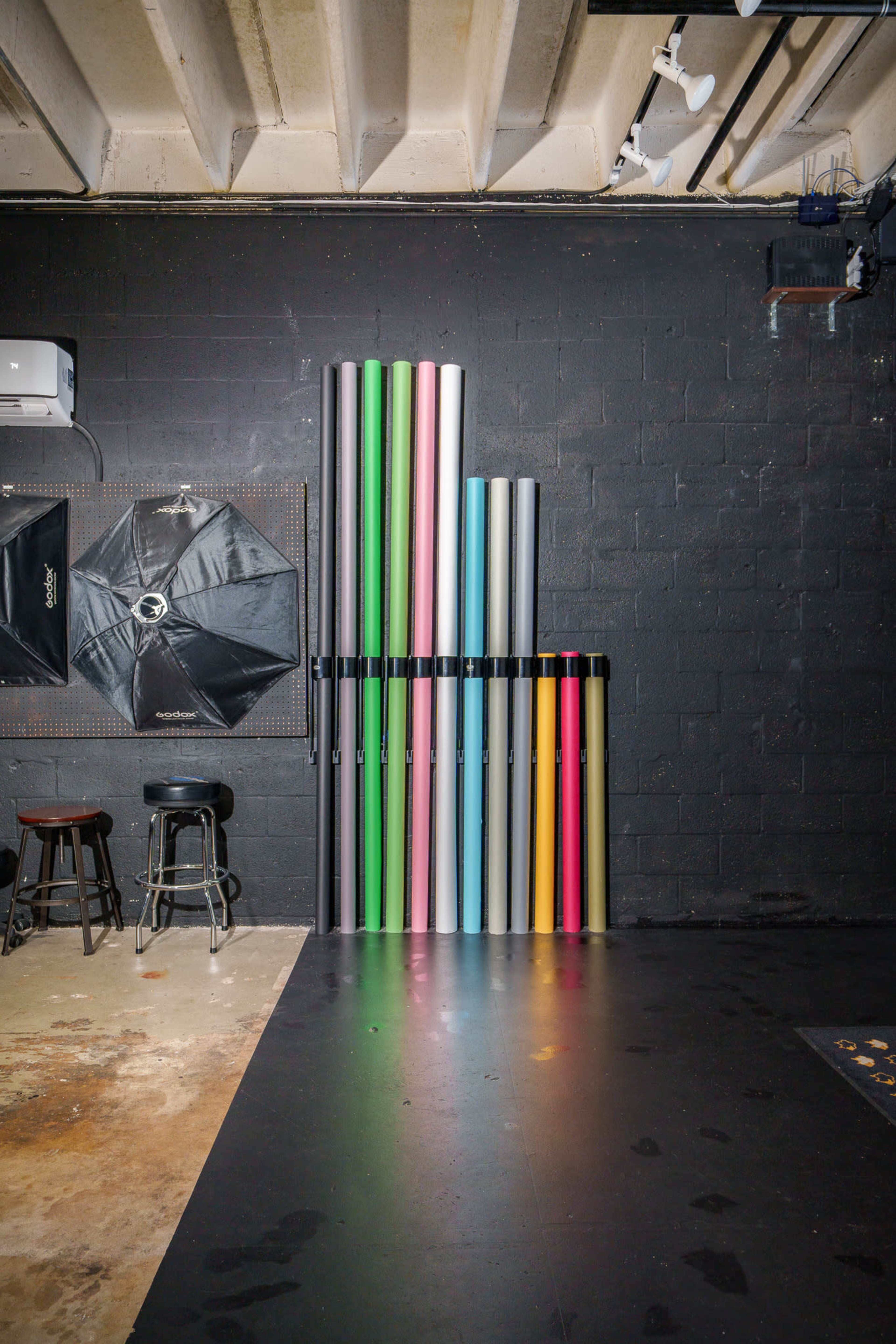 A collection of large rolls of seamless backdrop paper stands against a black wall in a studio, with a photography umbrella and two bar stools in the background.
