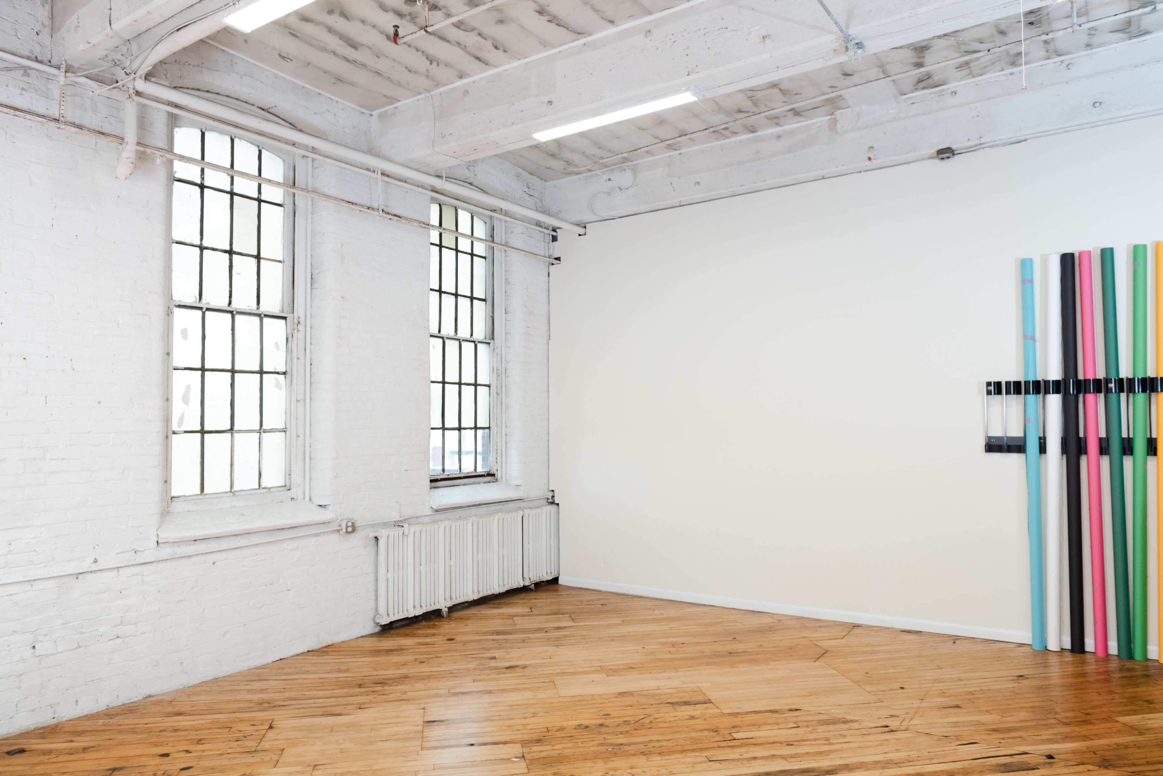 An empty room with white walls, wooden flooring, large windows, and a colorful artwork on one wall.
