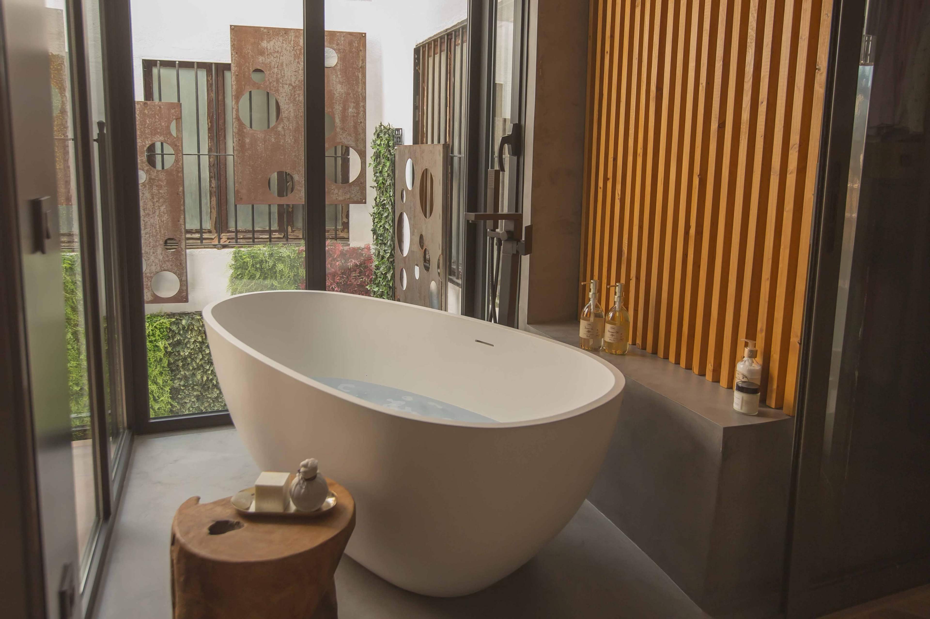 A modern bathroom featuring a freestanding white bathtub beside a brown wooden stool, with large windows providing a view of outdoor greenery.
