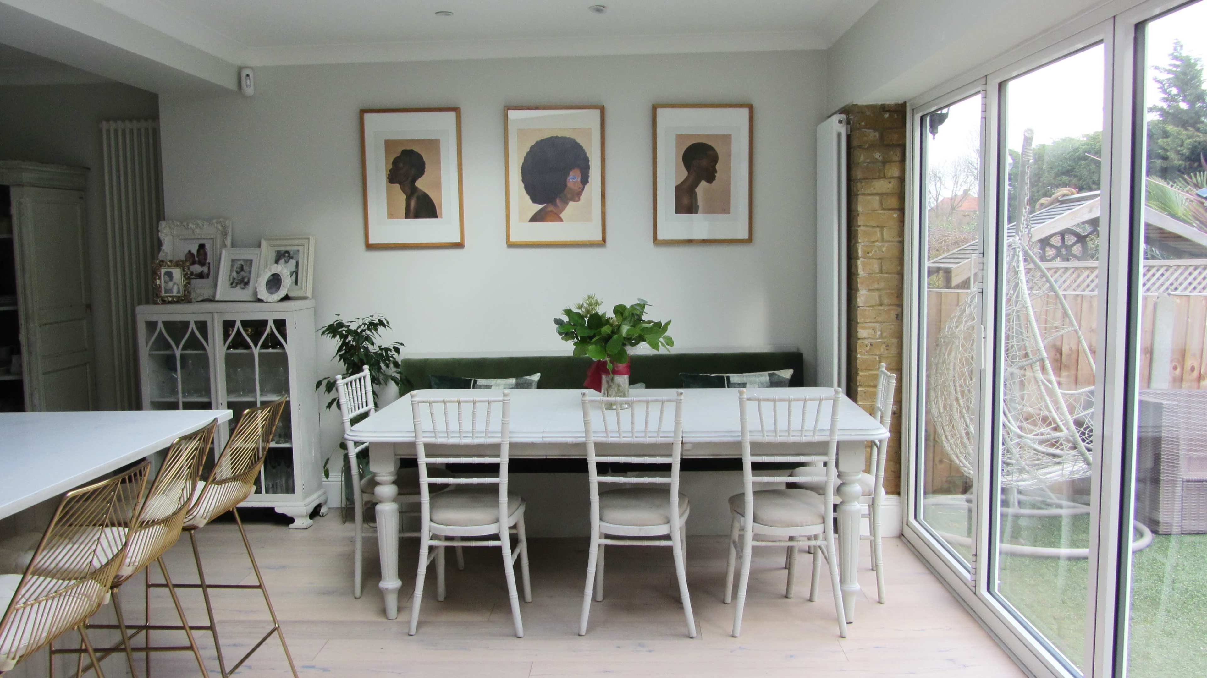 A dining area features a long white table surrounded by several white chairs, with framed portraits on the wall and a green banquette along one side.