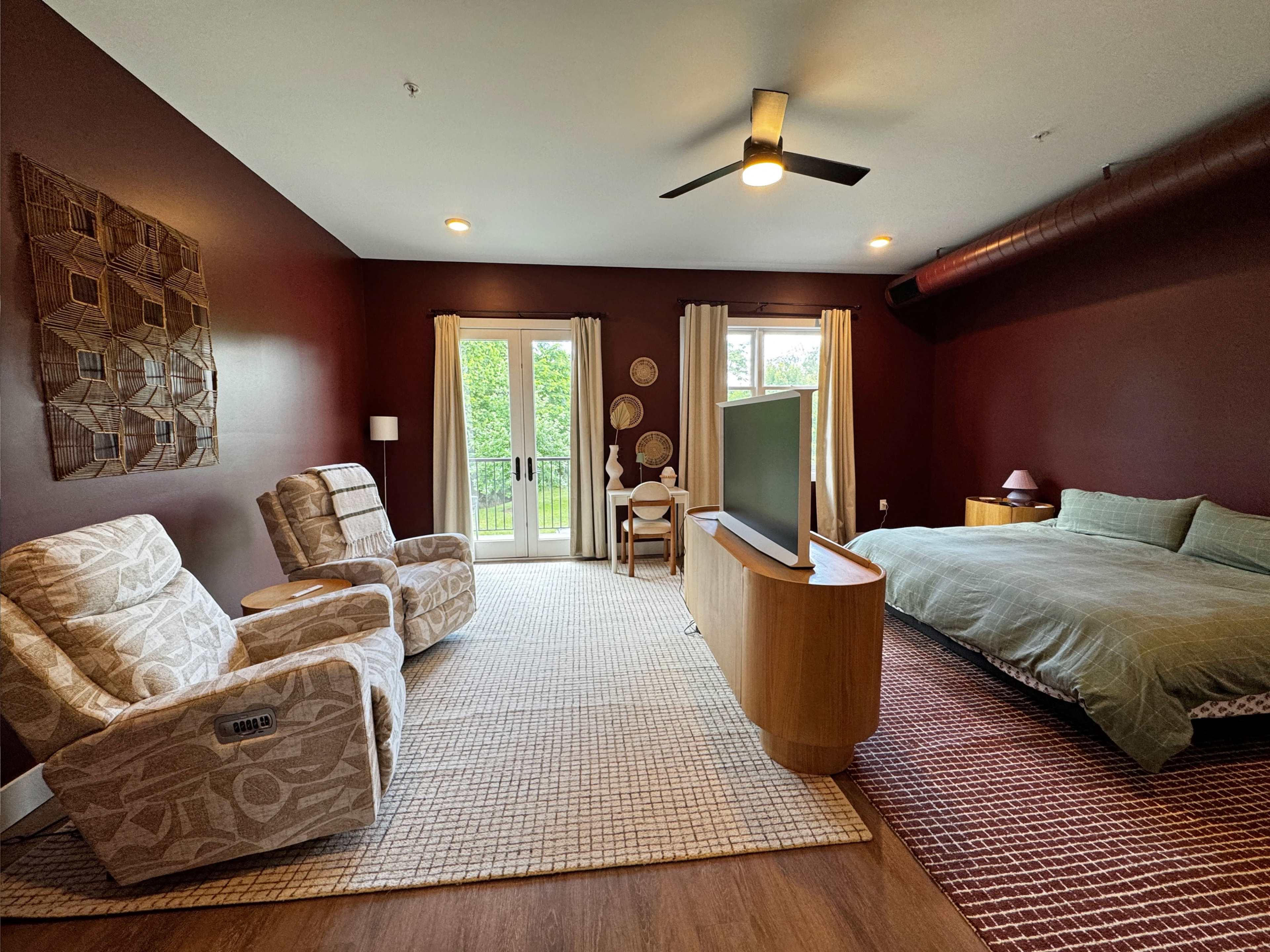 The image shows a modern bedroom featuring a large bed, two armchairs, a television mounted on a wooden console, and sliding glass doors leading to an outdoor area.