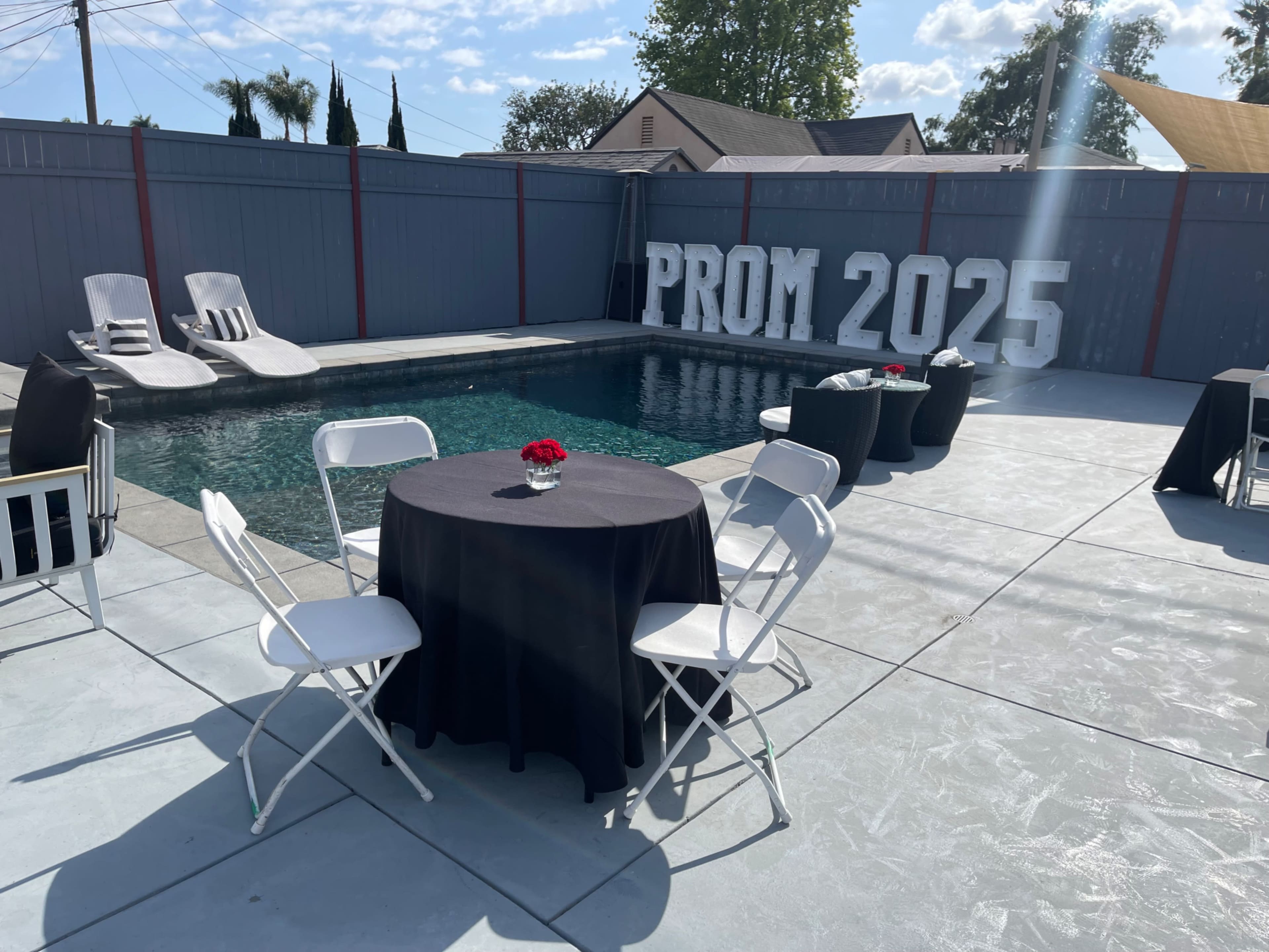 A decorated outdoor area features a swimming pool, tables with black tablecloths, and large letters spelling "PROM 2025" along a gray fence.