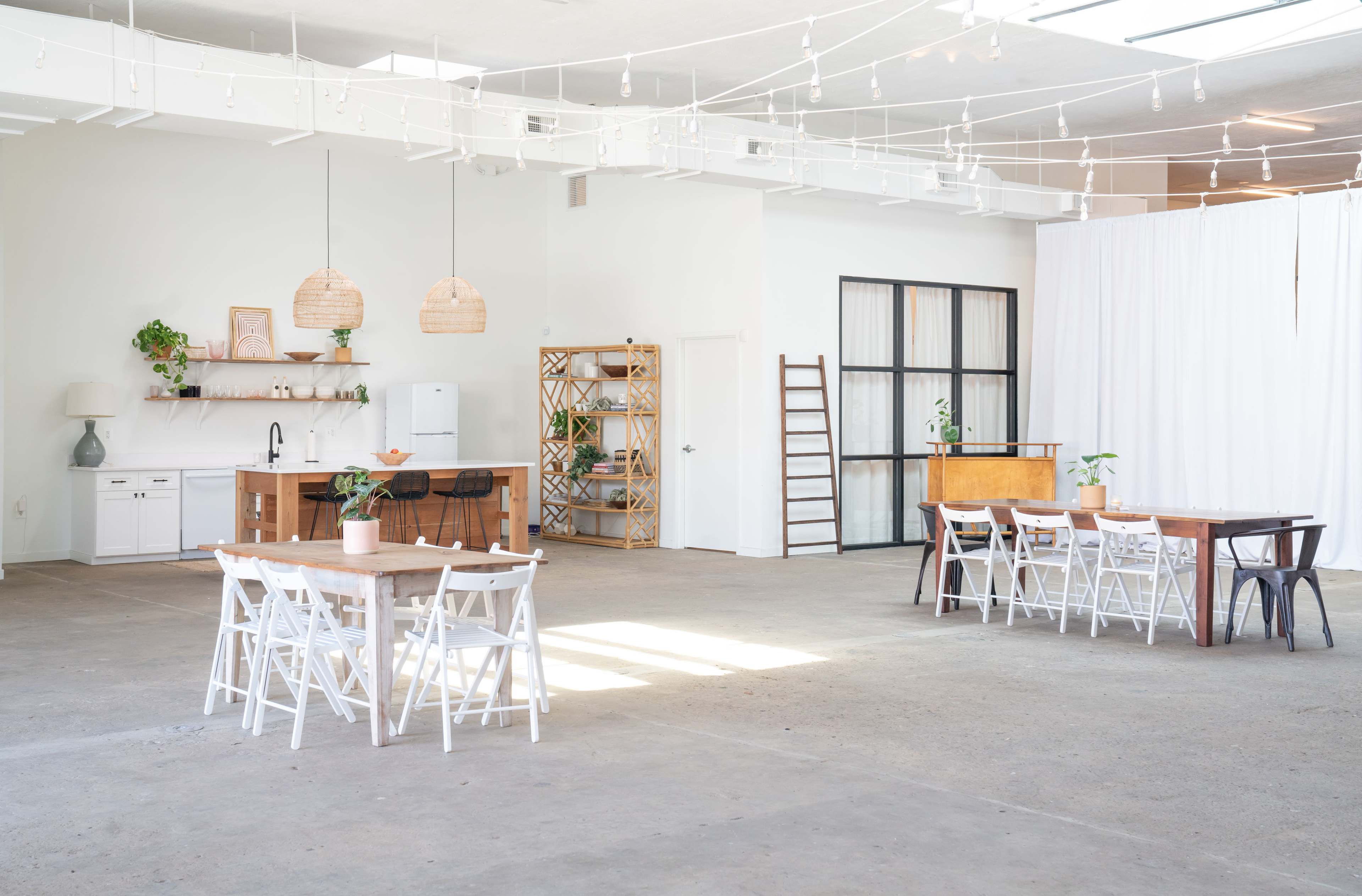 A spacious, well-lit room with wooden tables and chairs, a kitchen area, and decorative elements such as plants and hanging lights.