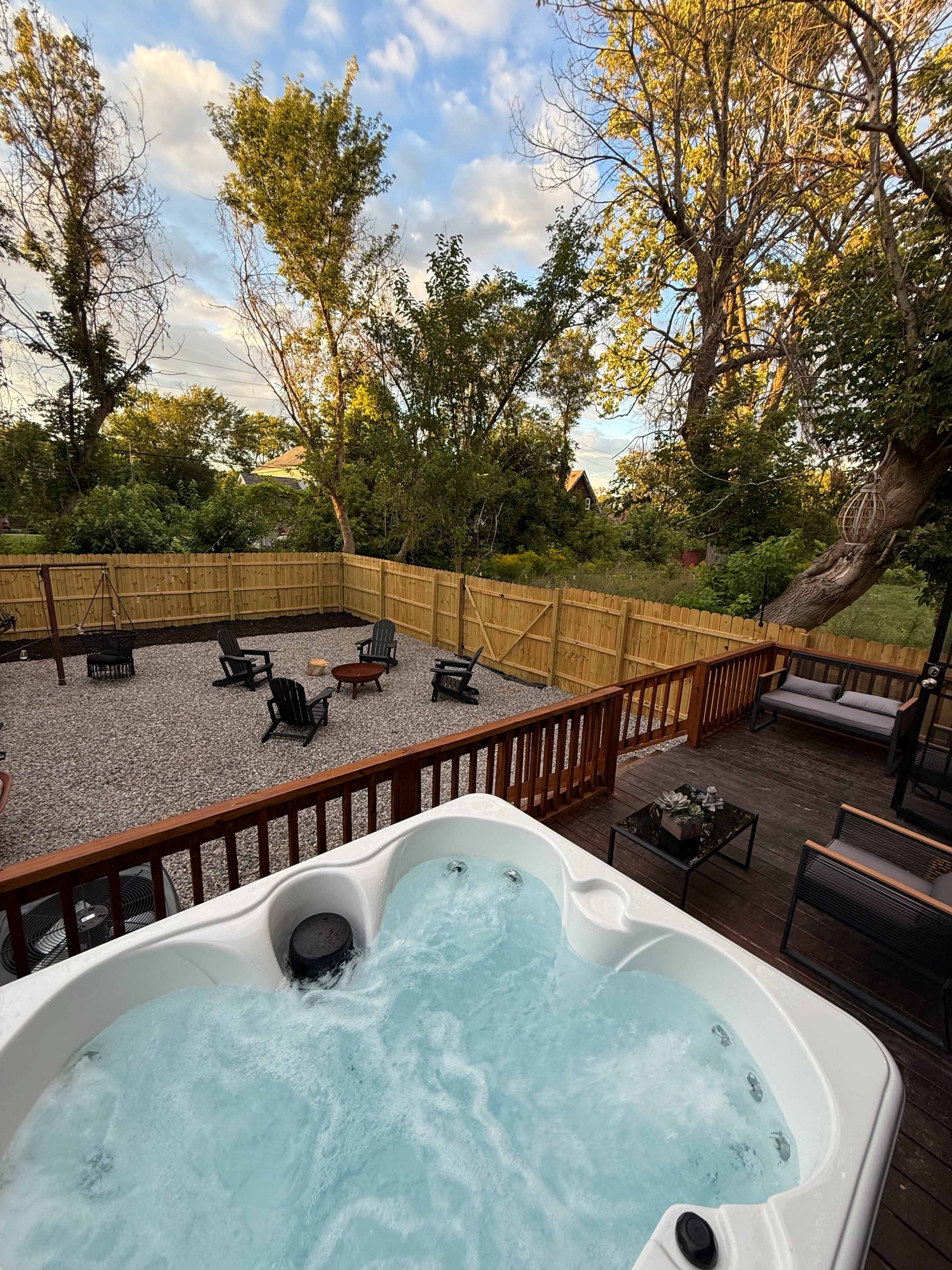 A hot tub is positioned on a wooden deck overlooking a spacious, fenced backyard with seating and a fire pit.