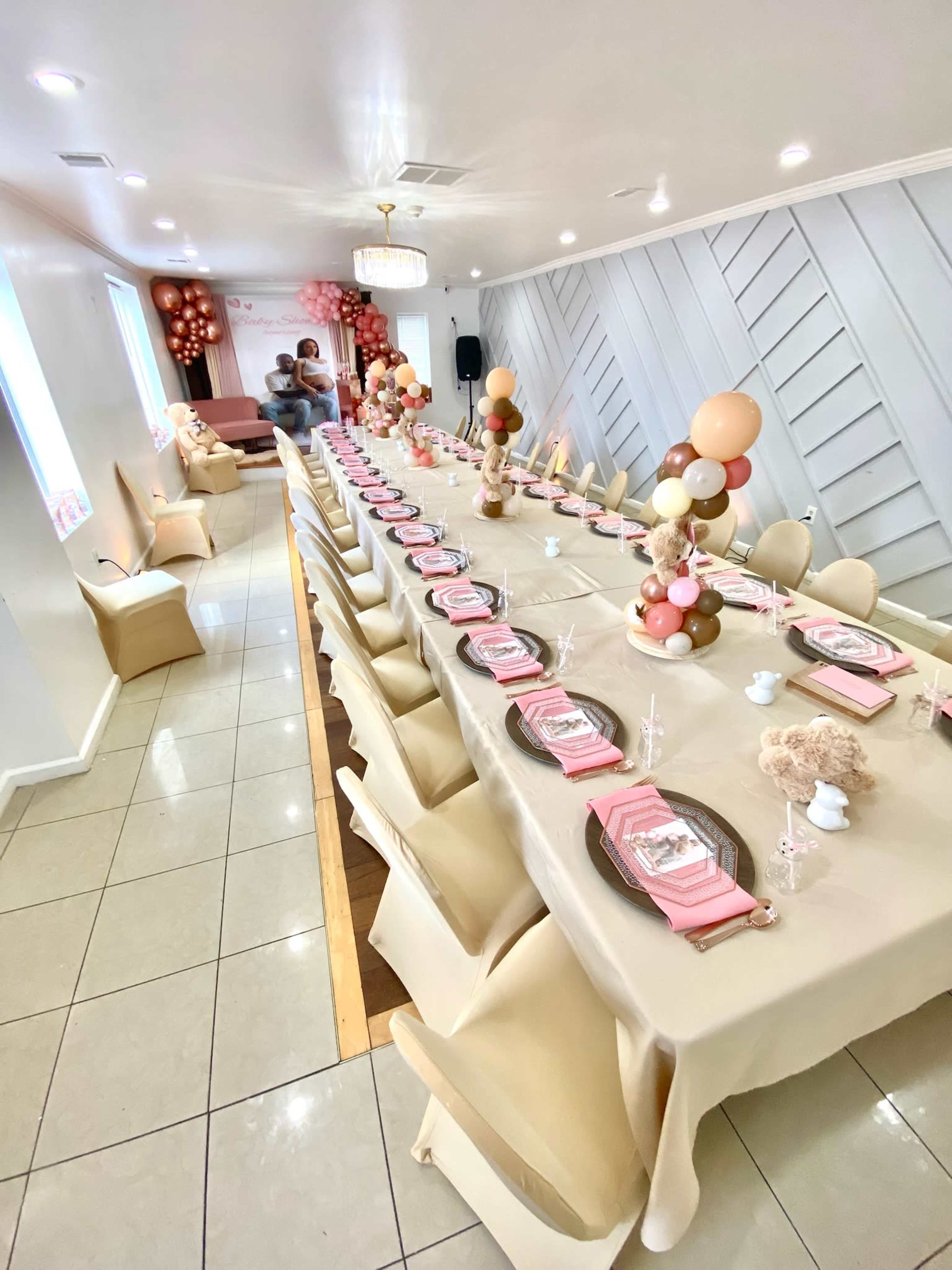 A long table is set for a celebration, adorned with beige and pink tablecloths, decorative balloons, and place settings, in a well-lit indoor space.