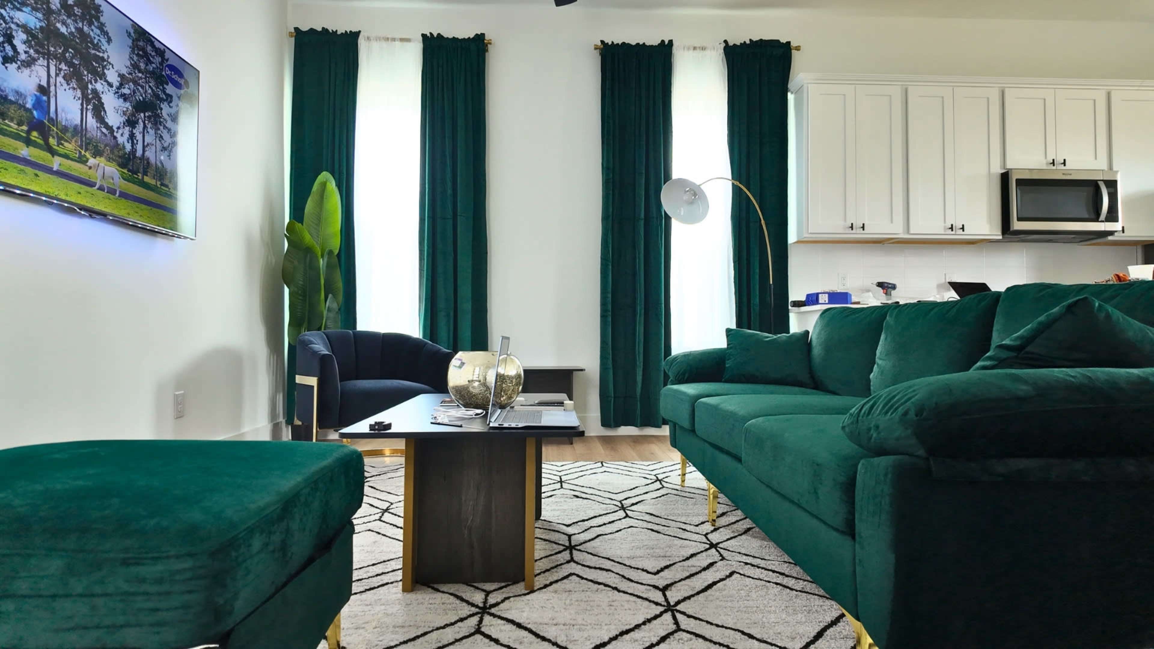 The image shows a modern living room featuring a green velvet sofa, a dark blue armchair, a coffee table, and large windows with white curtains.