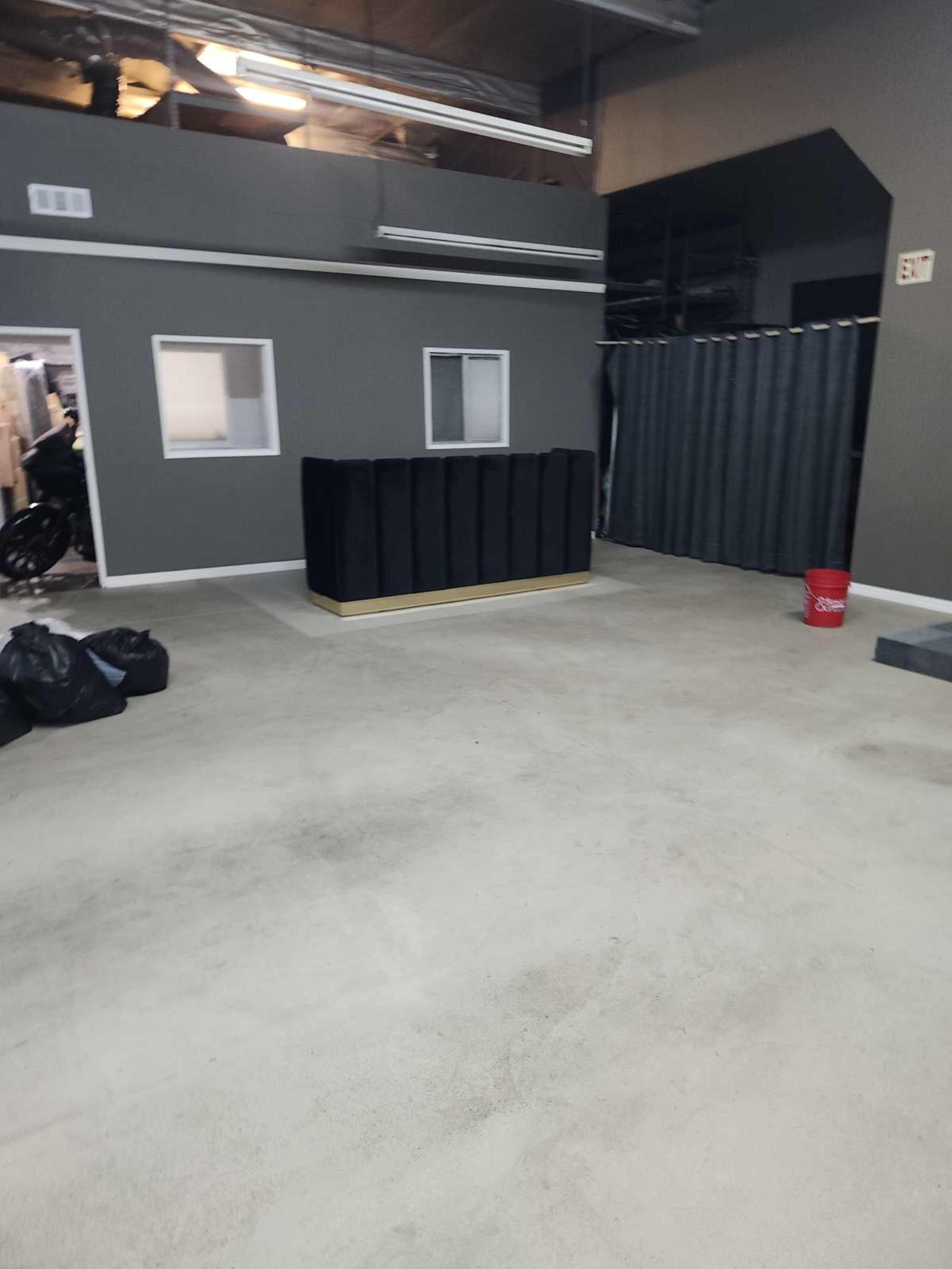 The image shows a large, empty room with a gray wall, a black curtain, a counter at the center, and bags along the floor.
