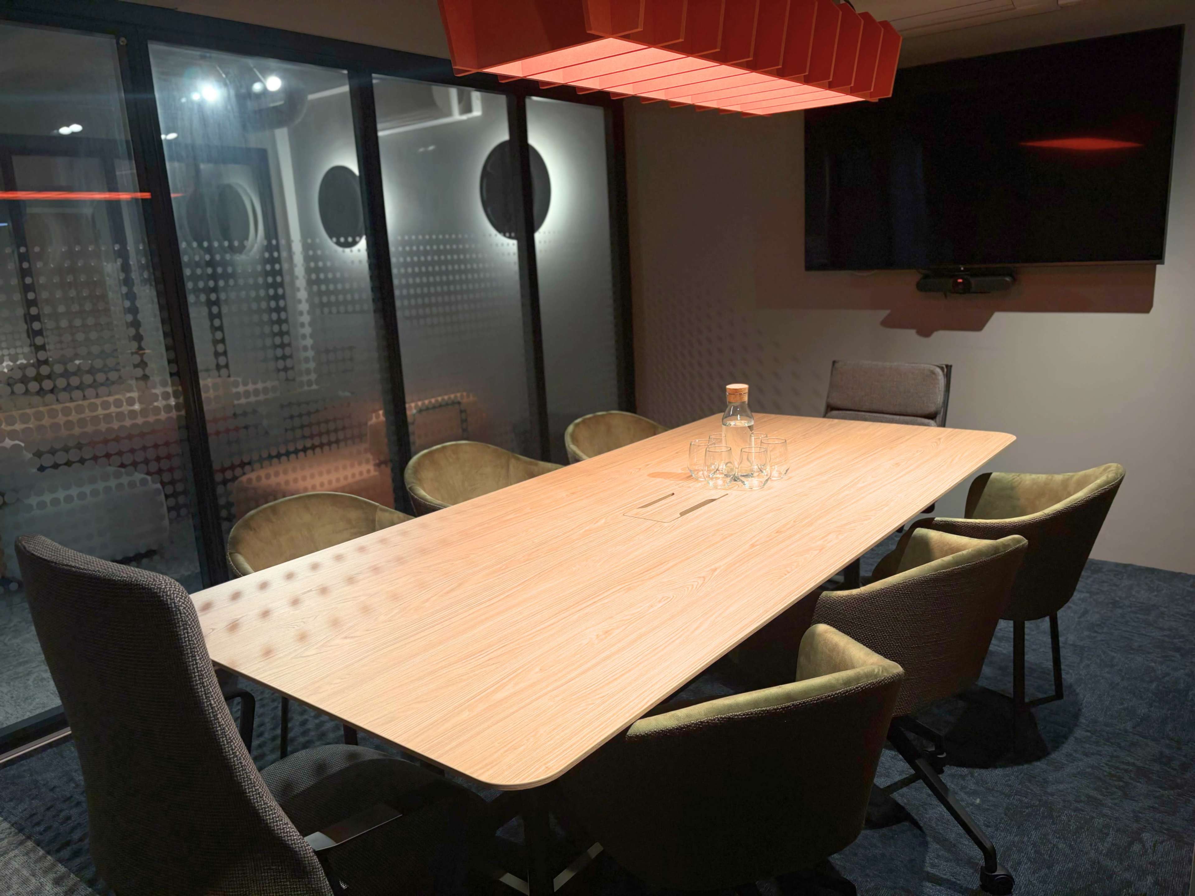 A modern conference room features a long wooden table surrounded by six chairs, with a large television screen mounted on the wall and a glass wall providing visibility to the outside.
