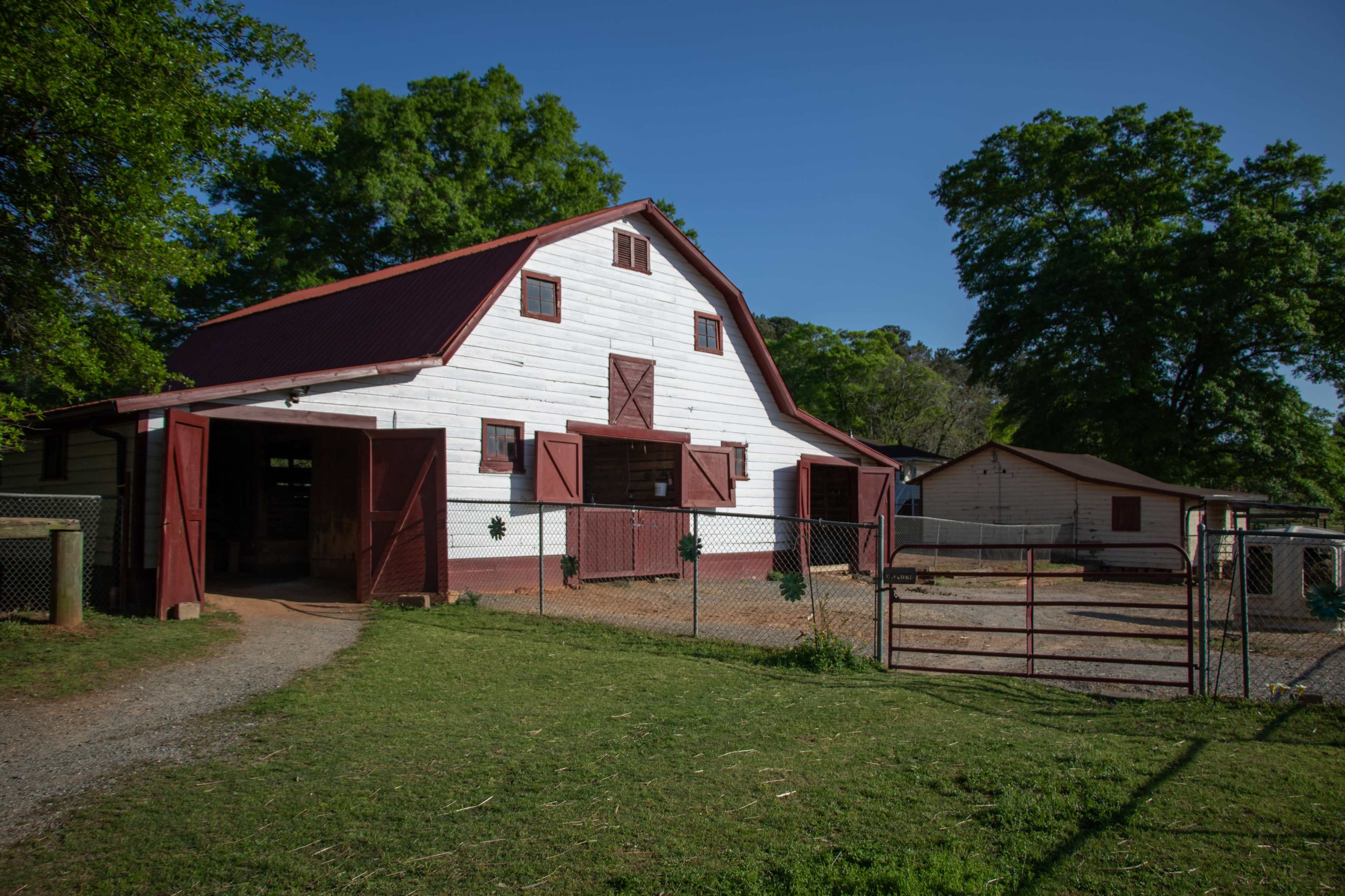 The image shows a white barn with a red roof and doors, surrounded by a fenced area and green trees nearby.