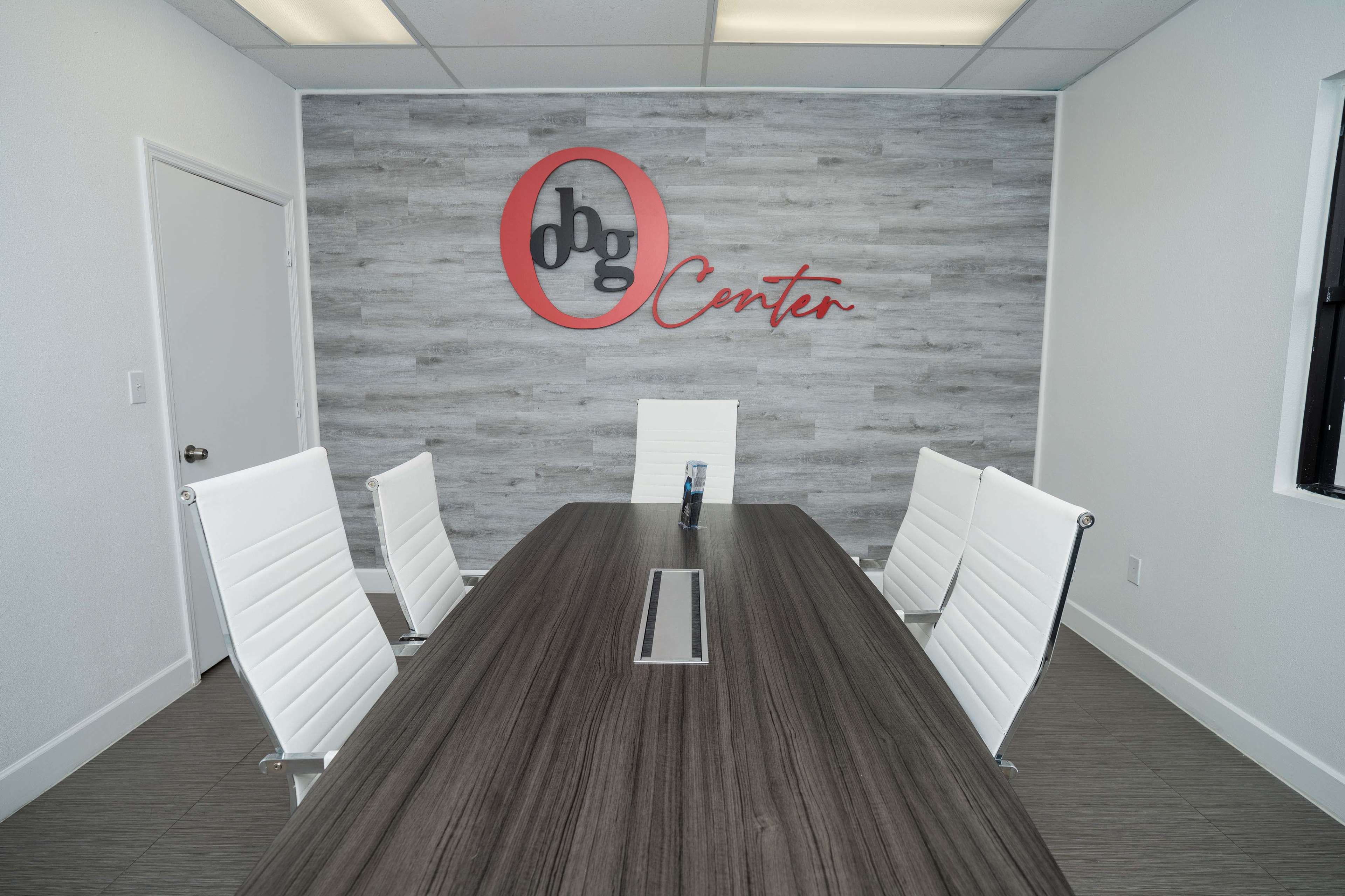 A conference room features a wooden table with a central cable management slot, surrounded by four white chairs and a branded wall with the text "dbg Center."