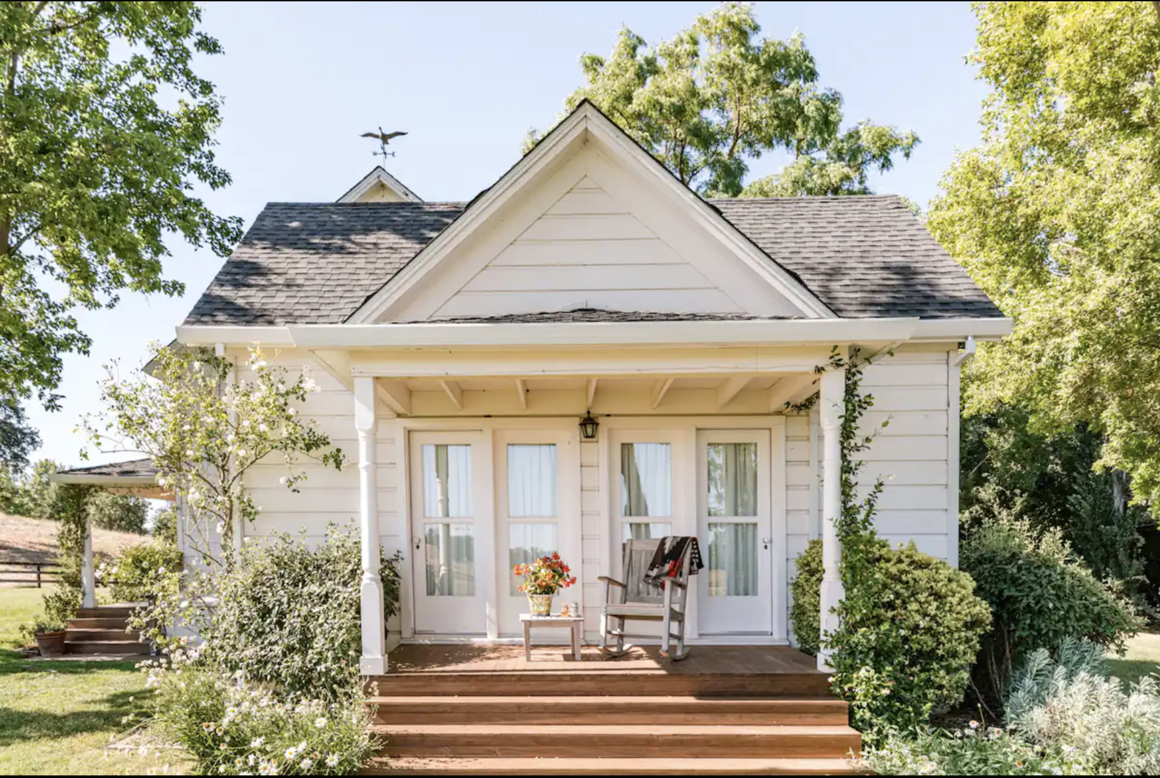 A quaint white cottage with a front porch features double doors, a rocking chair, and surrounding greenery.