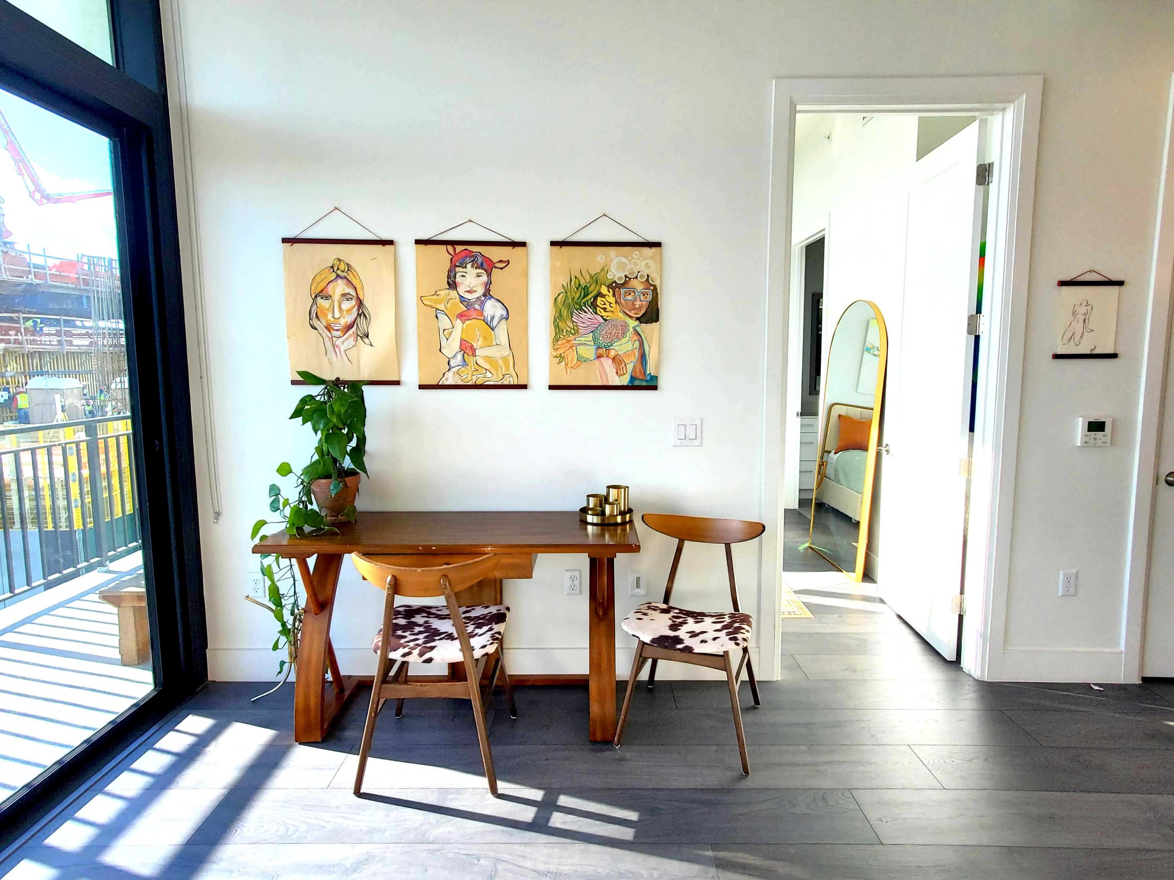 A small dining area featuring a wooden table and two chairs, with three art pieces hanging on the wall and a doorway leading to another room.