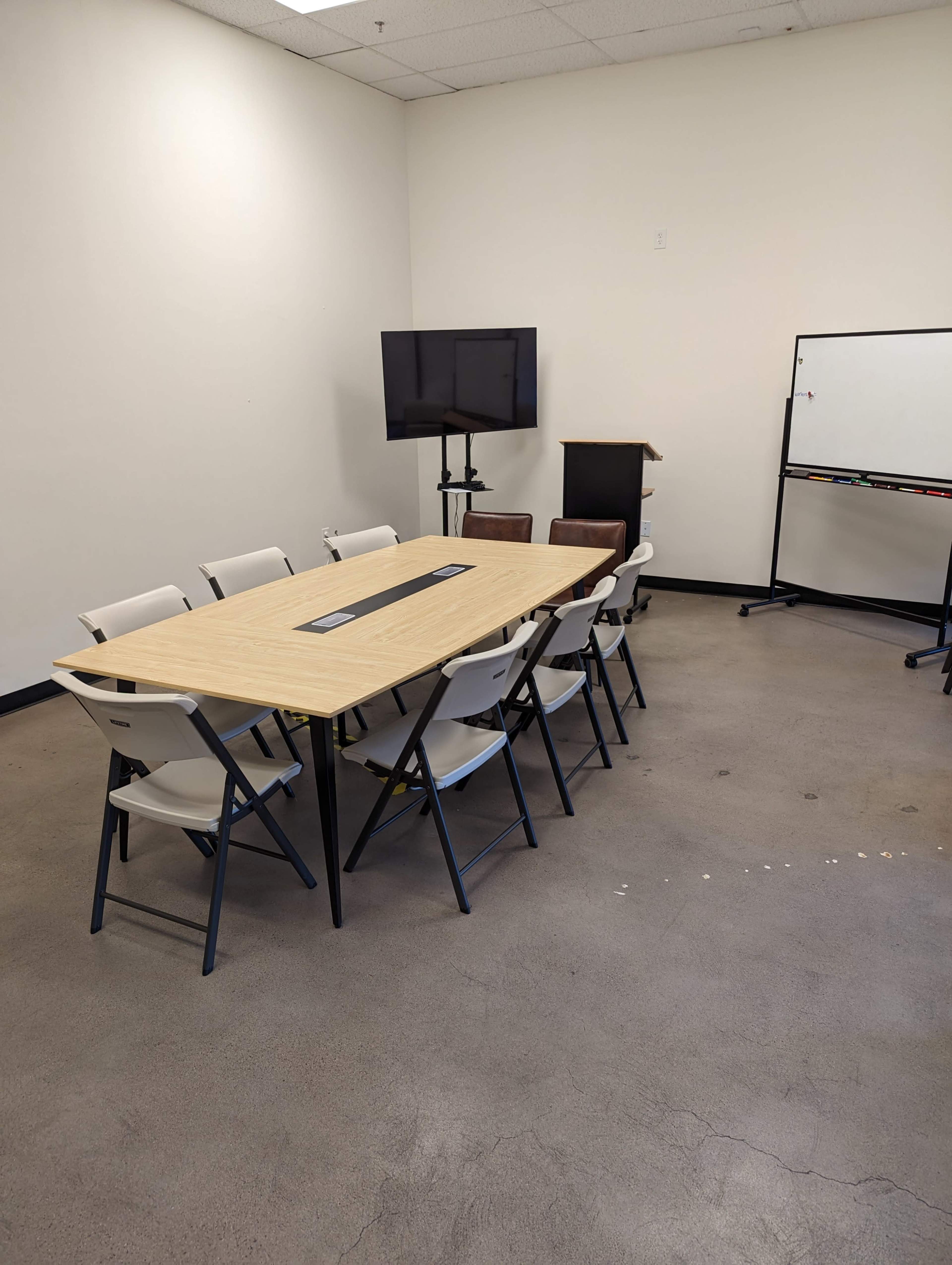 A conference room features a long wooden table surrounded by folding chairs, a television on a stand, a podium, and a whiteboard.