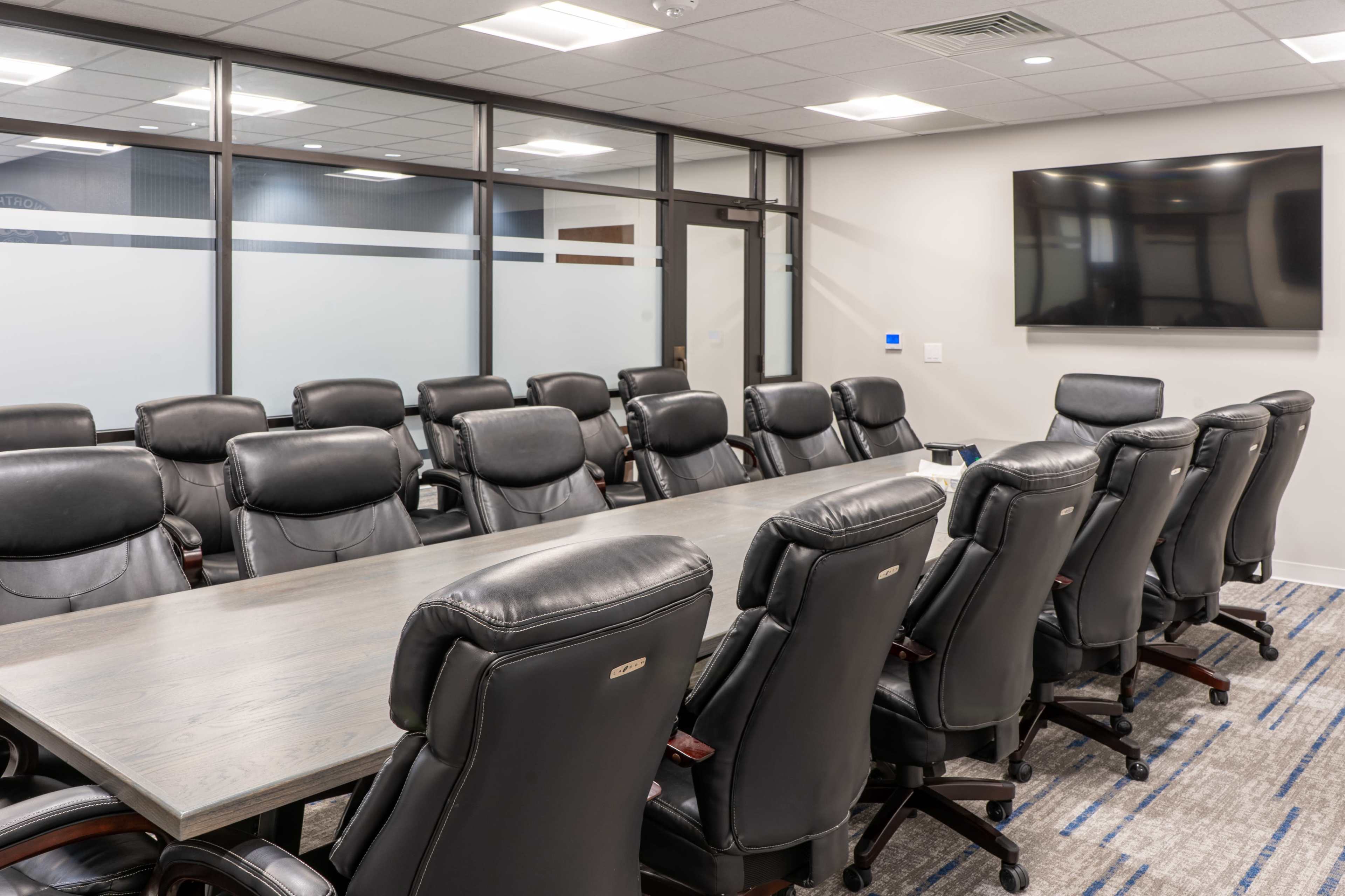 A long conference table with black leather chairs is set up in a modern meeting room featuring a wall-mounted television and glazed glass partitions.