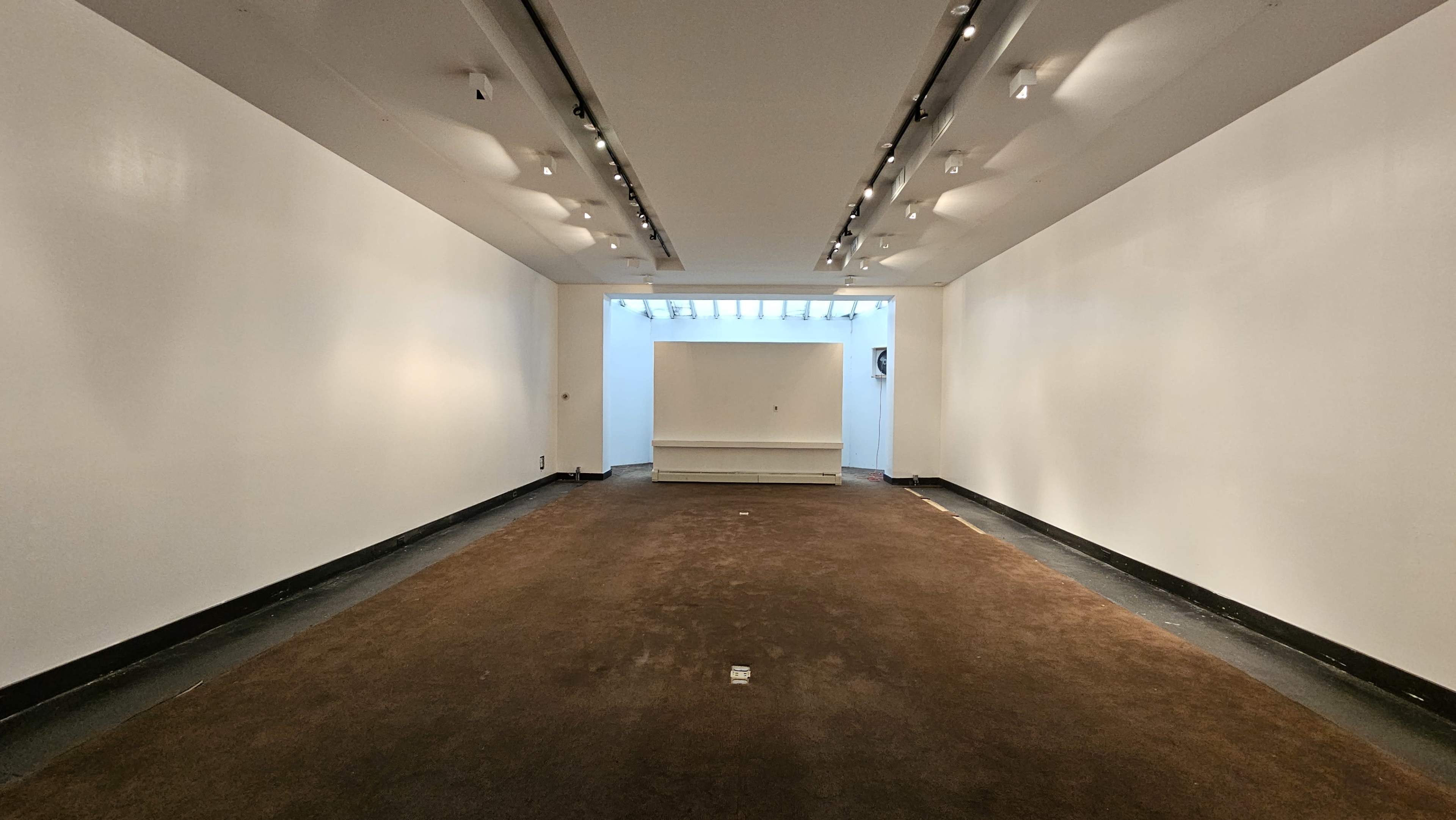 The image shows a spacious, empty gallery with white walls, a dark carpeted floor, and a bright opening at the far end.