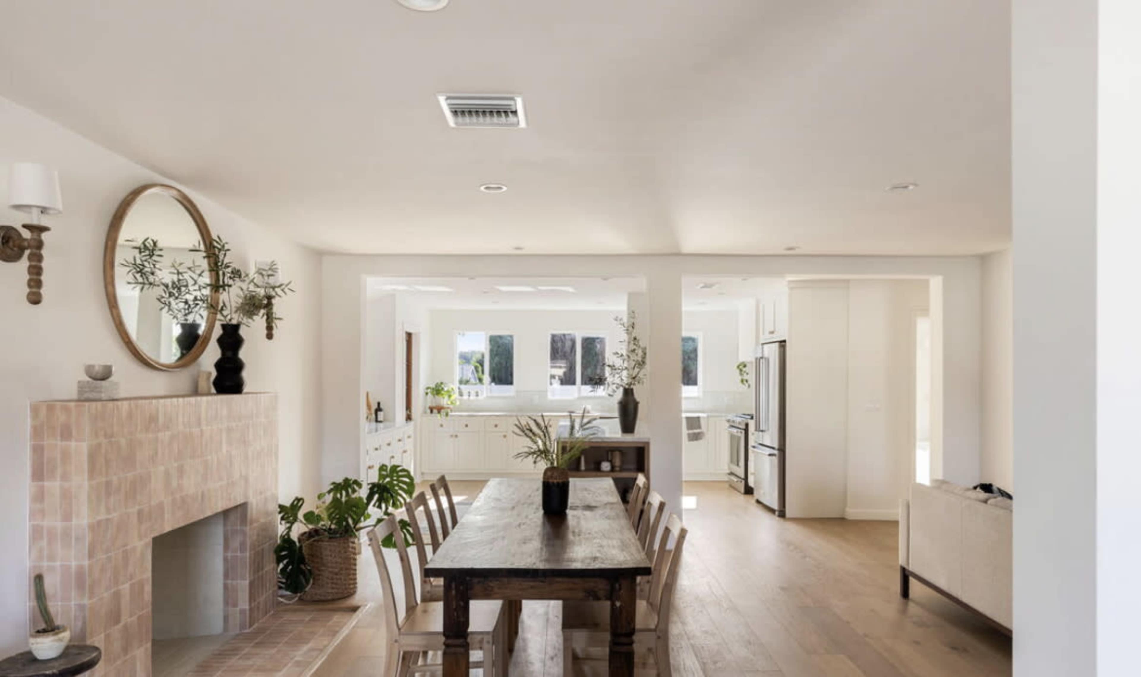The image shows a modern kitchen and dining area with a wooden table, a fireplace, and large windows allowing natural light.