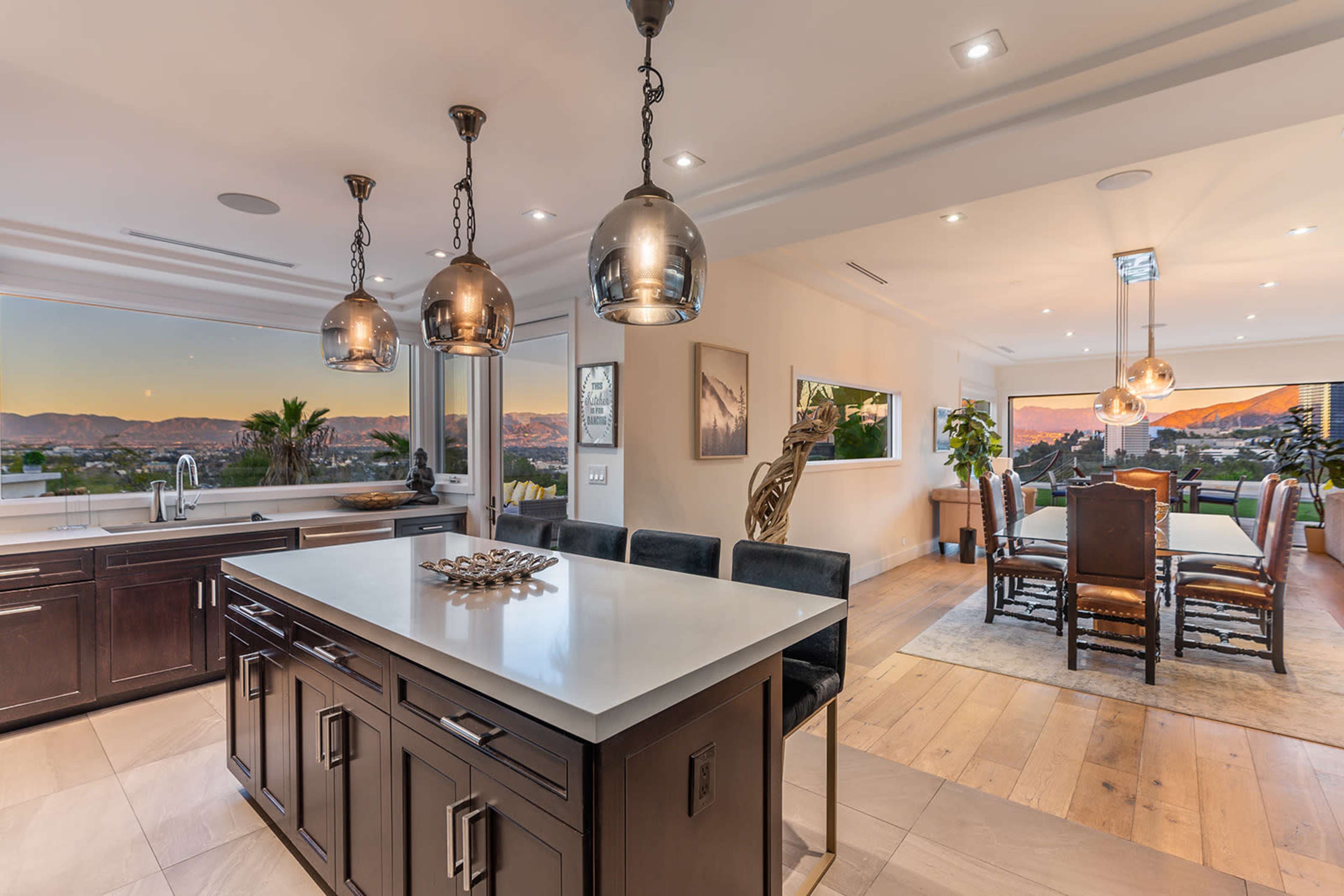 A modern kitchen with a central island and pendant lighting overlooks a dining area and a scenic view of the mountains through large windows.