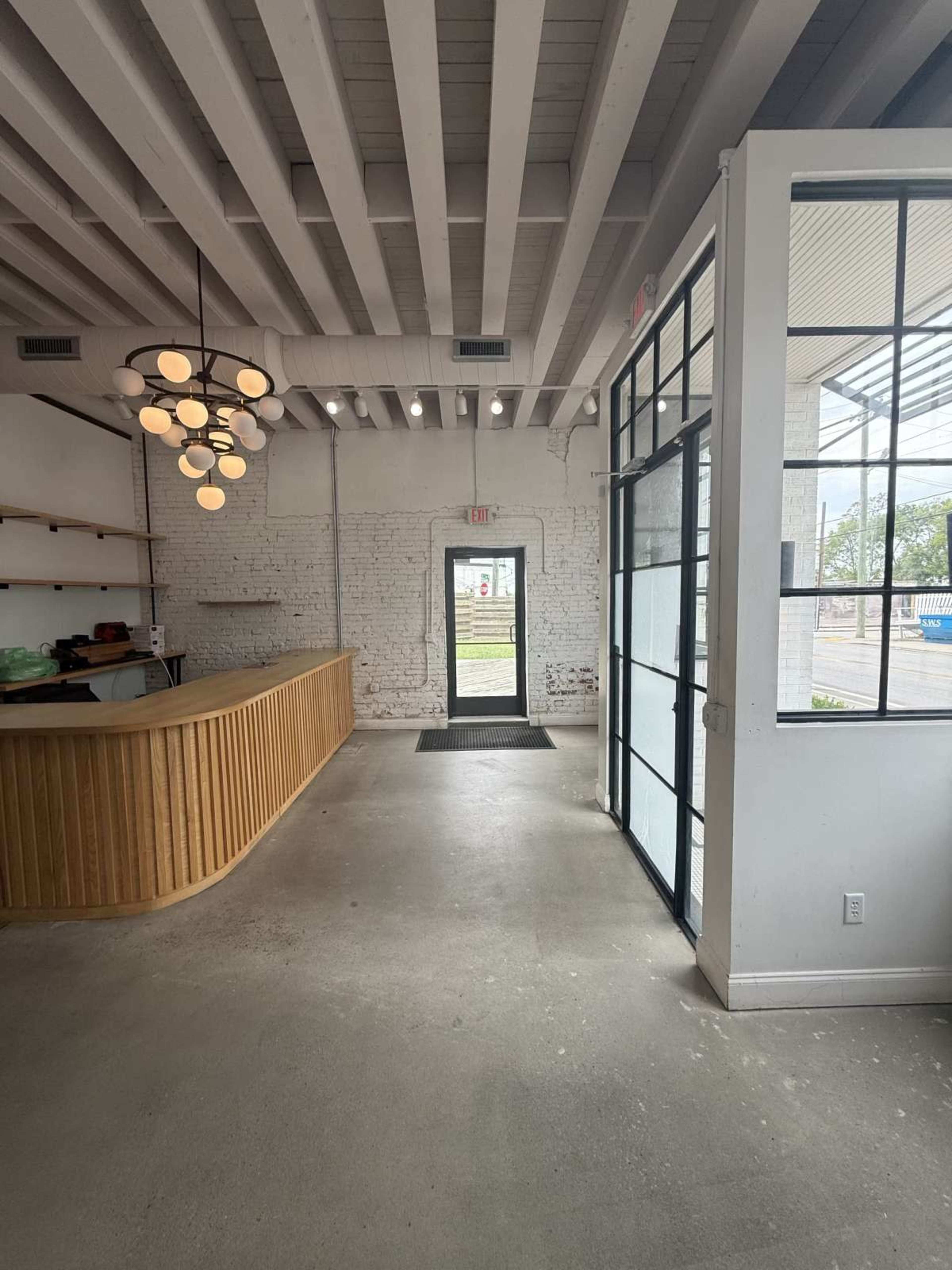The image shows a spacious, modern interior of a commercial space with a wooden reception desk, exposed beams, and a glass door leading outside.